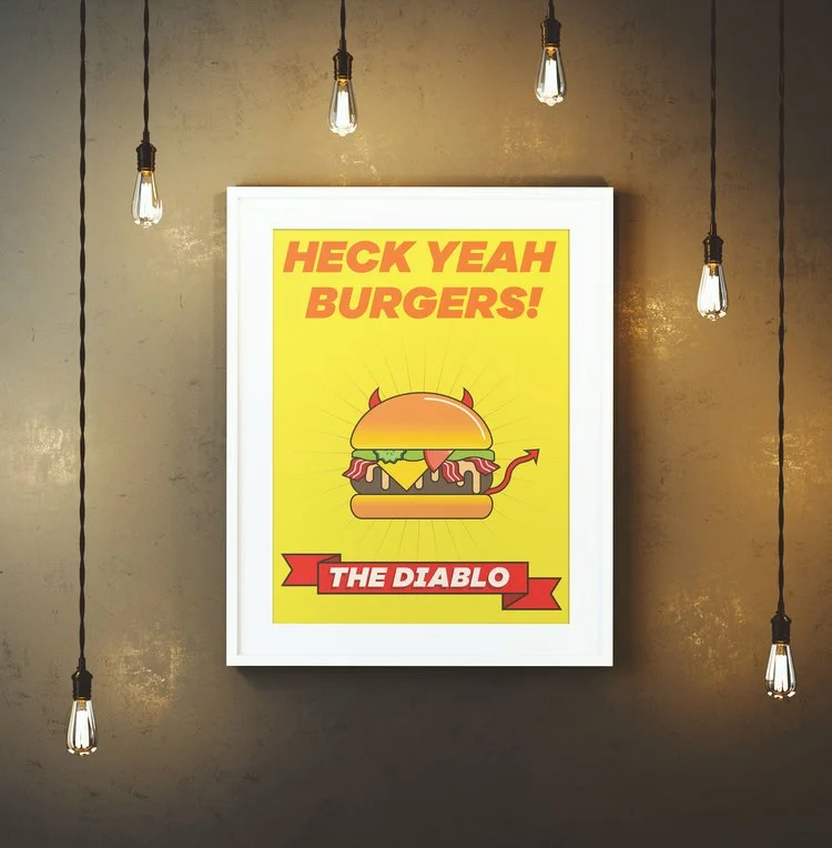 A framed poster of a cartoon burger with devil horns and tail on a yellow background. Bold text above says 'HECK YEAH BURGERS!' and a red banner below reads 'THE DIABLO'. Five hanging light bulbs illuminate the poster against a textured beige wall.