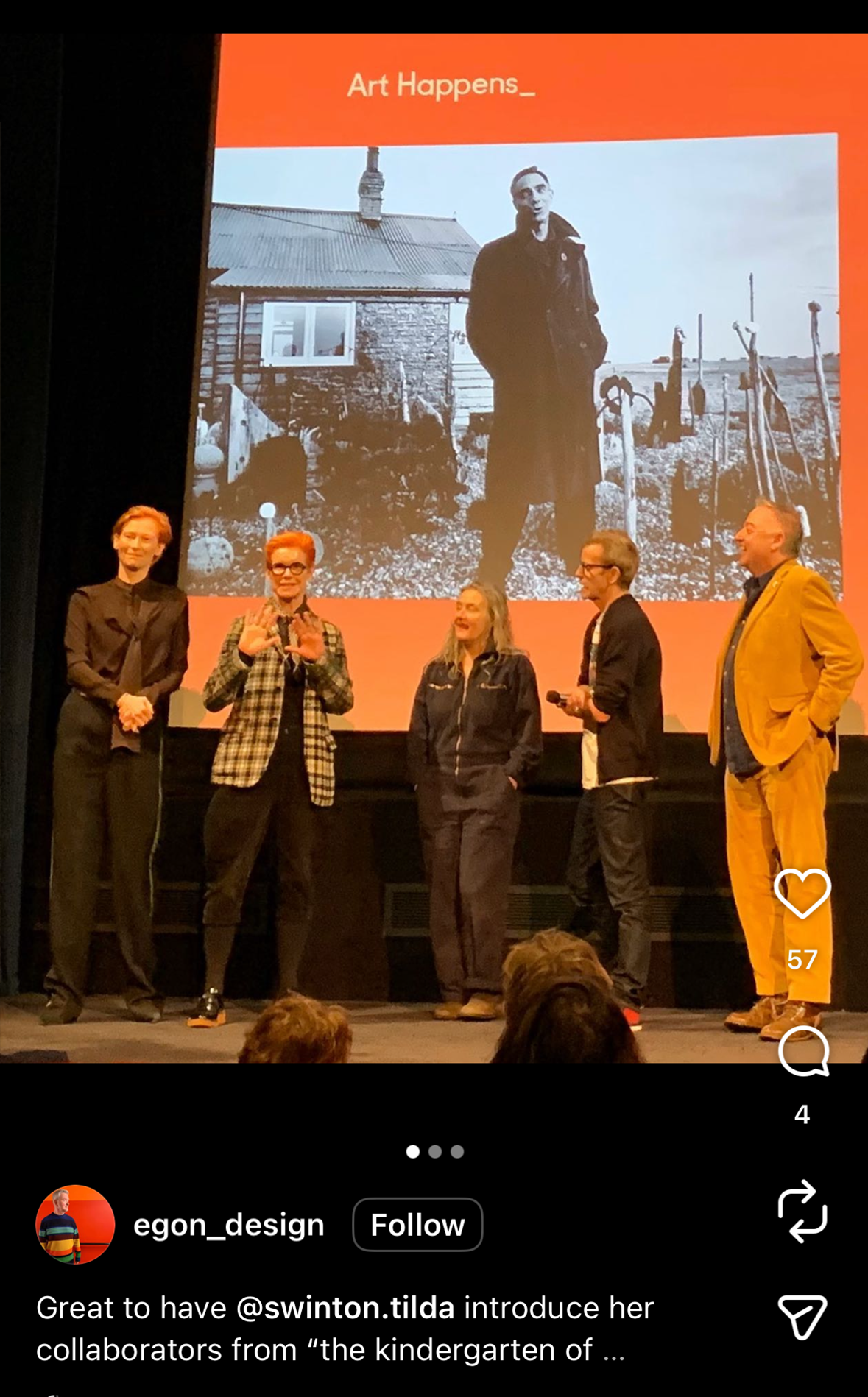 Five individuals stand on a stage in front of a large screen showing a black-and-white photo of a person standing outside a rustic house with tools in the background. The stage has a dark backdrop, and the people are engaged, likely in a presentation