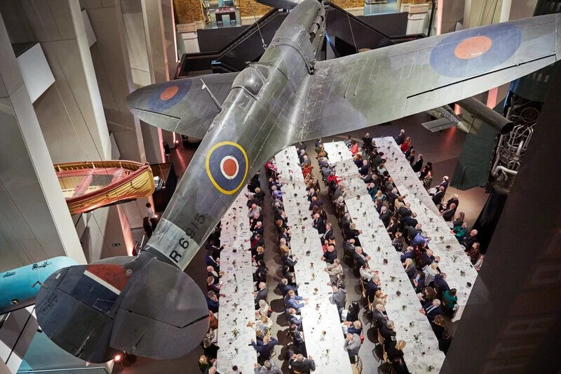 An aerial view of a large indoor museum with a suspended vintage military aircraft overhead and a crowd of people seated at long tables.