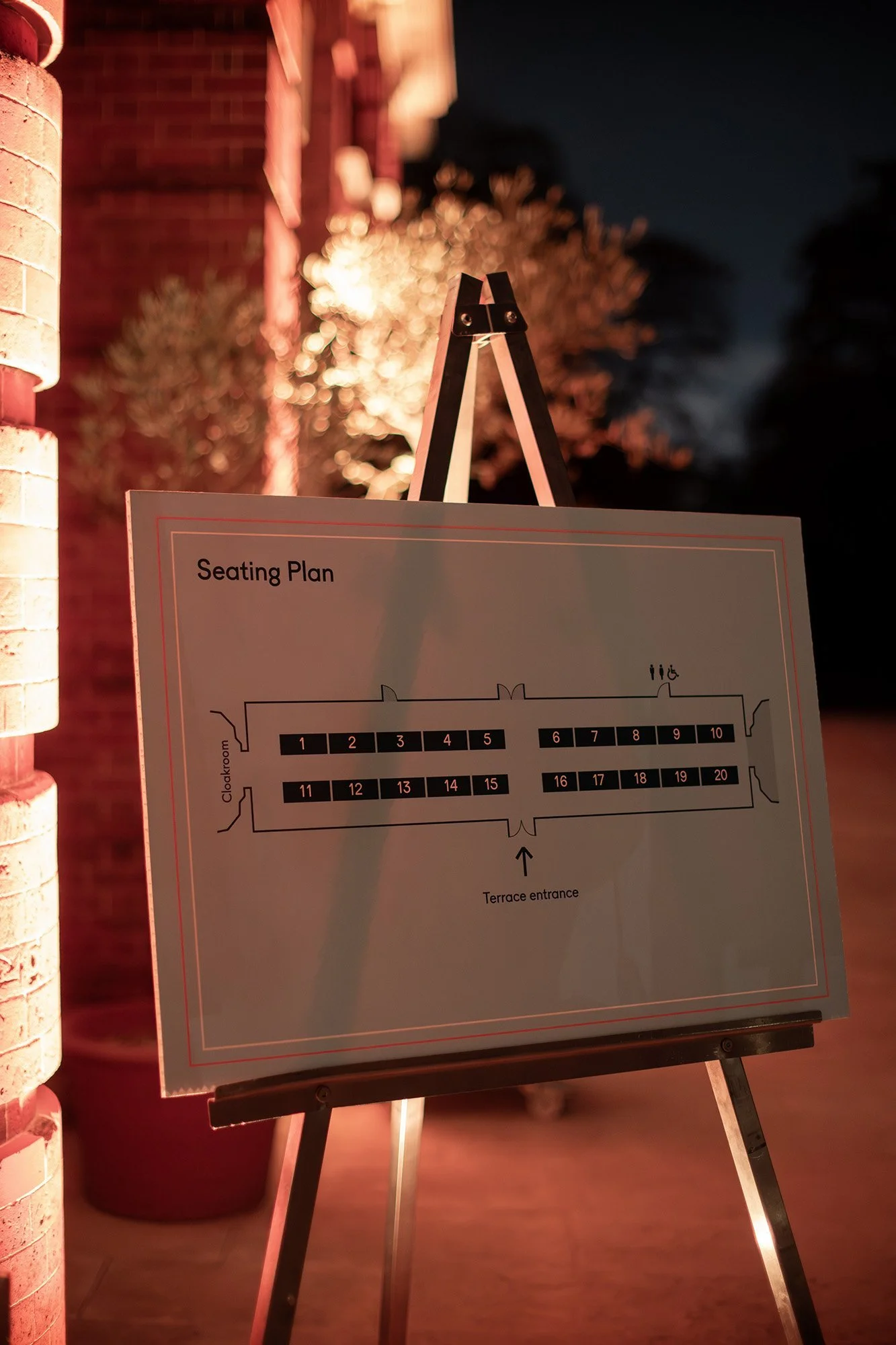 Nighttime outdoor scene featuring a seating plan on an easel with a brick building wall on the left, illuminated by warm lighting, and blurred trees and dark sky in the background.