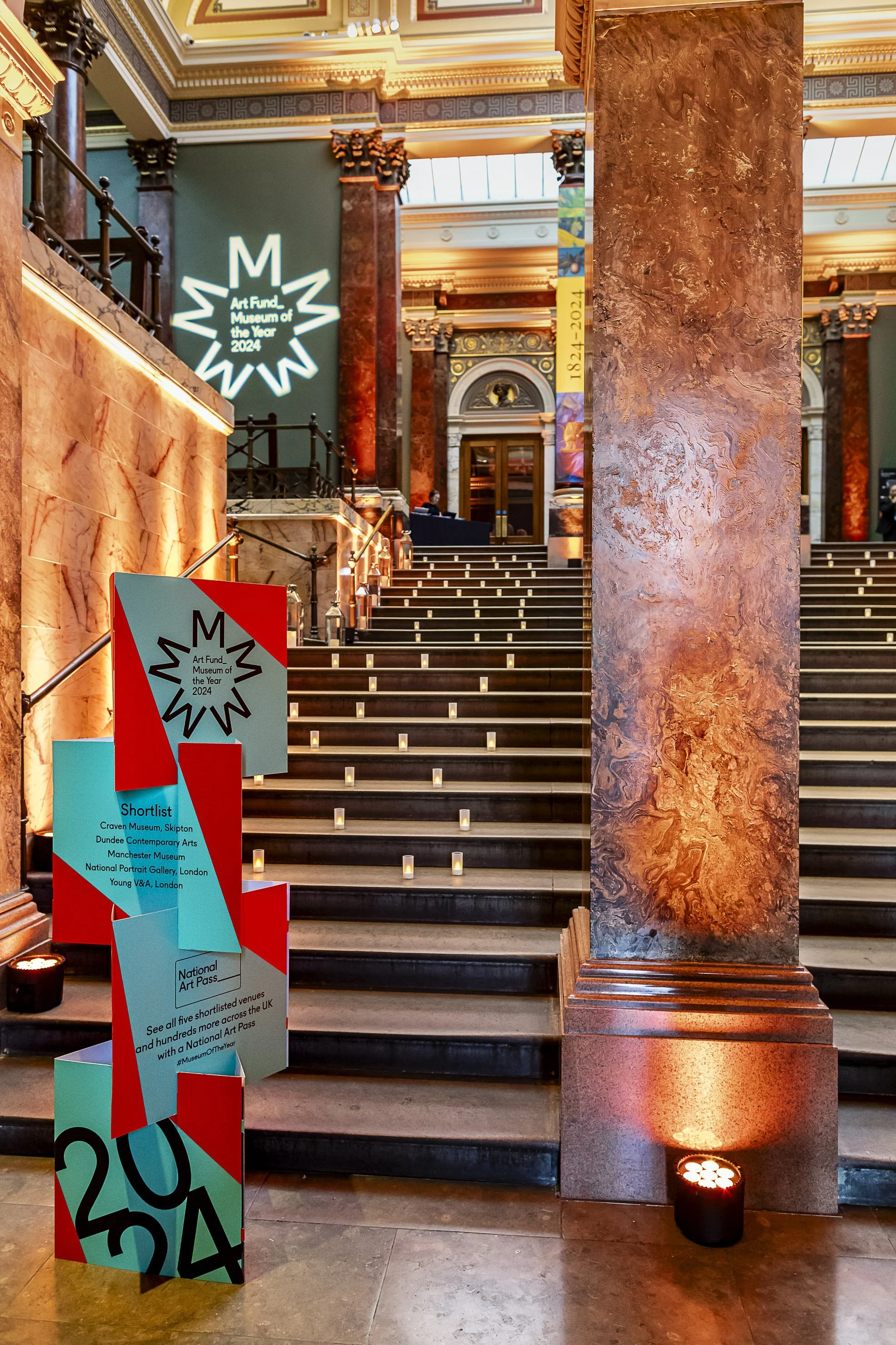 Interior of a grand historic building with marble columns, decorative moldings, and a staircase with candles along the sides. There are signs indicating the museum's shortlists and awards for 2024.
