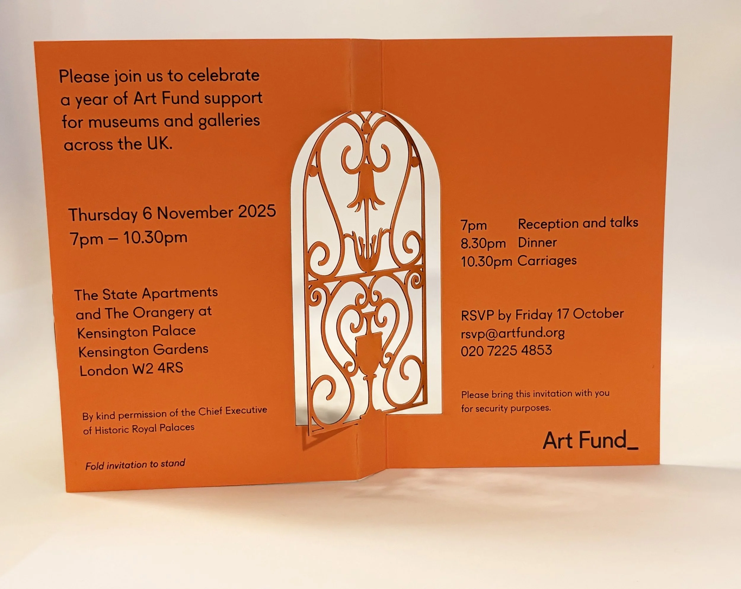 Open orange fold invitation with text for art fundraising event, featuring a decorative wrought iron gate illustration on the right side.
