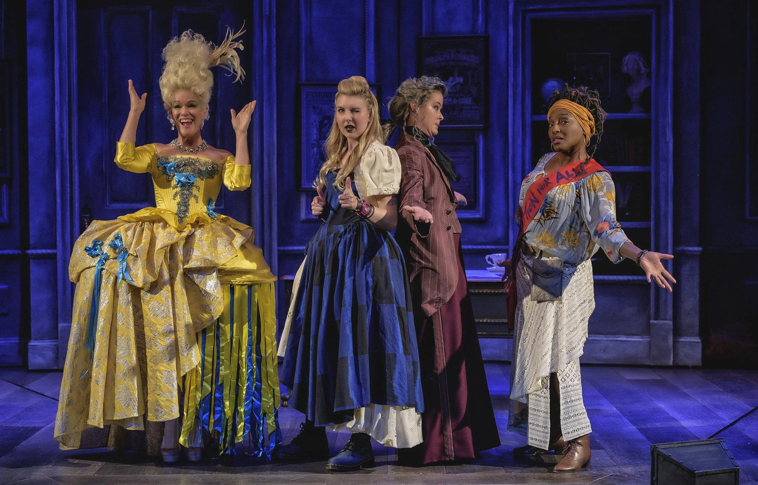 Four women in colorful costumes performing on a stage with blue-lit wooden walls in the background.