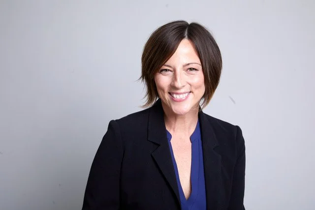 A woman with short dark hair smiling, wearing a black blazer and a blue blouse, standing against a plain light gray background.