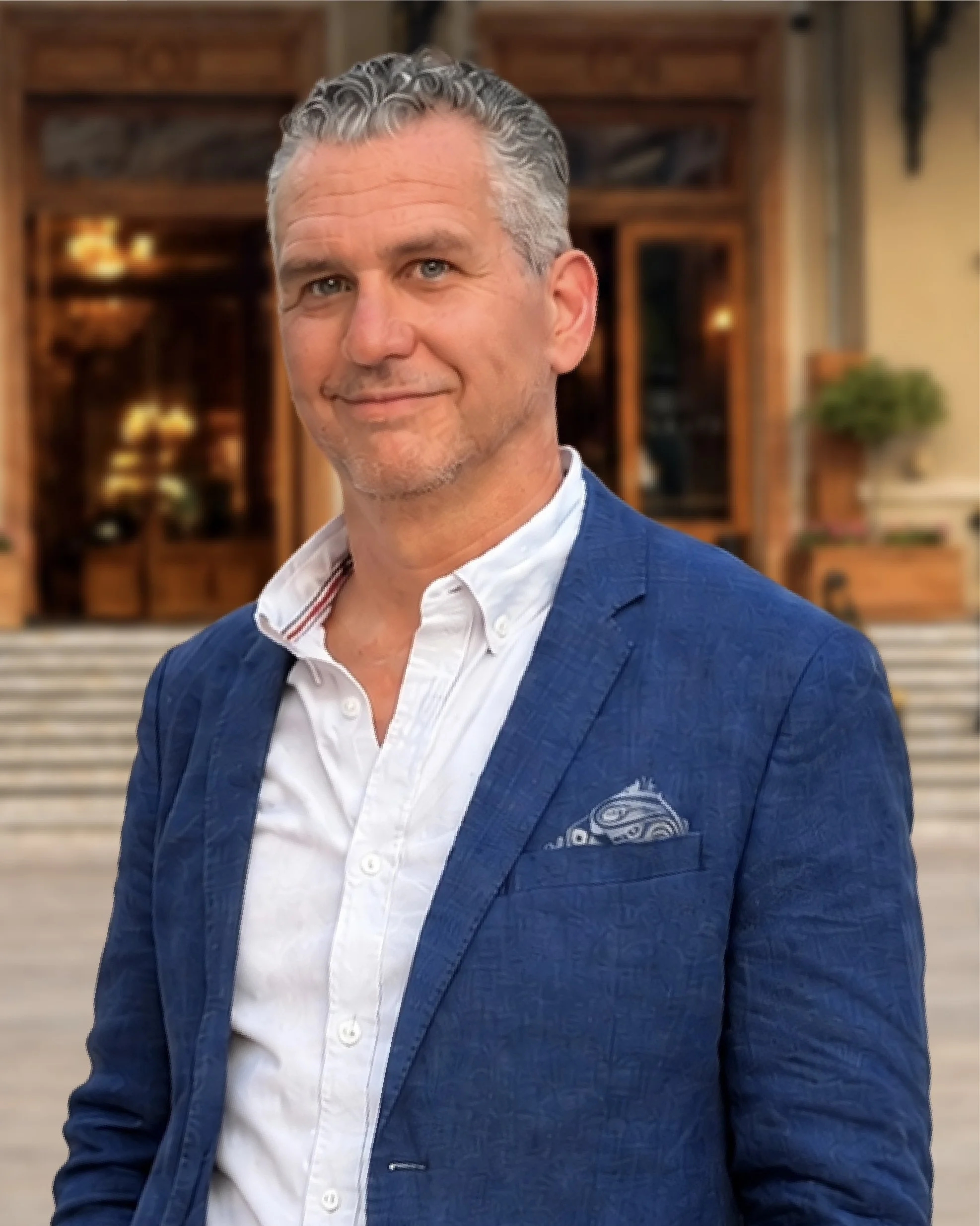 Portrait of a middle-aged man with gray hair and blue eyes, wearing a light blue collared shirt, smiling softly against a plain neutral background.