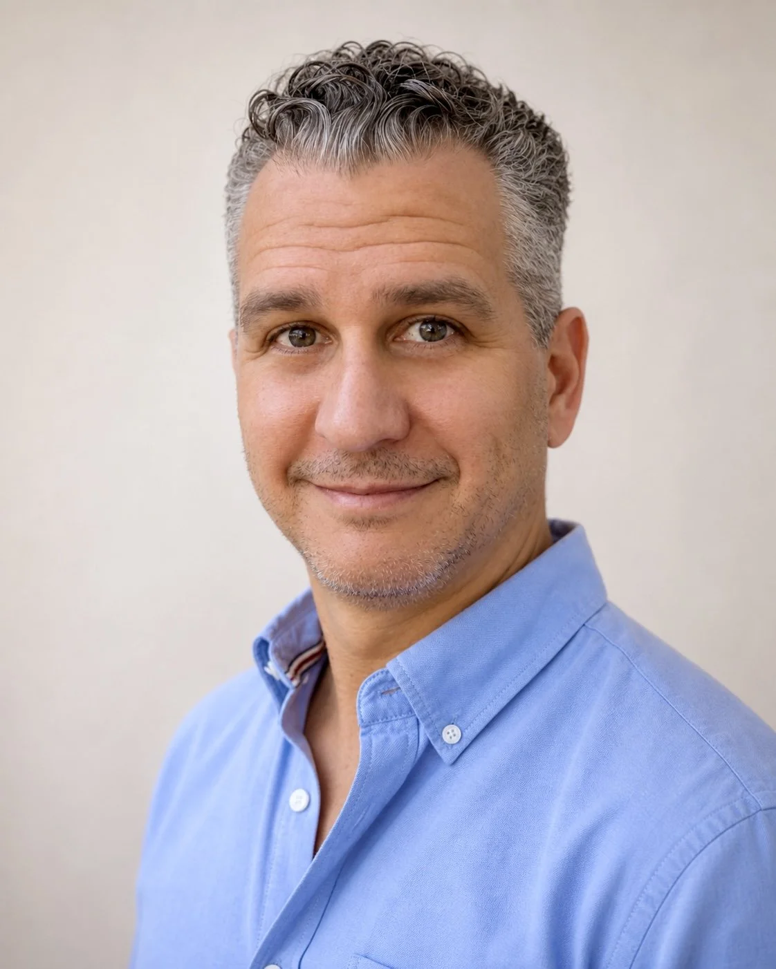 Portrait of a middle-aged man with gray hair and blue eyes, wearing a light blue collared shirt, smiling softly against a plain neutral background.