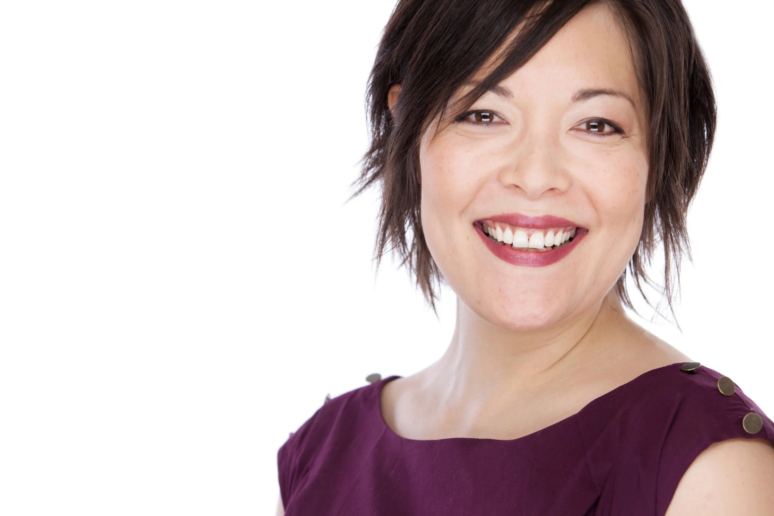 A woman with short dark hair smiling at the camera, wearing a purple top with gold button details on the shoulders, against a white background.