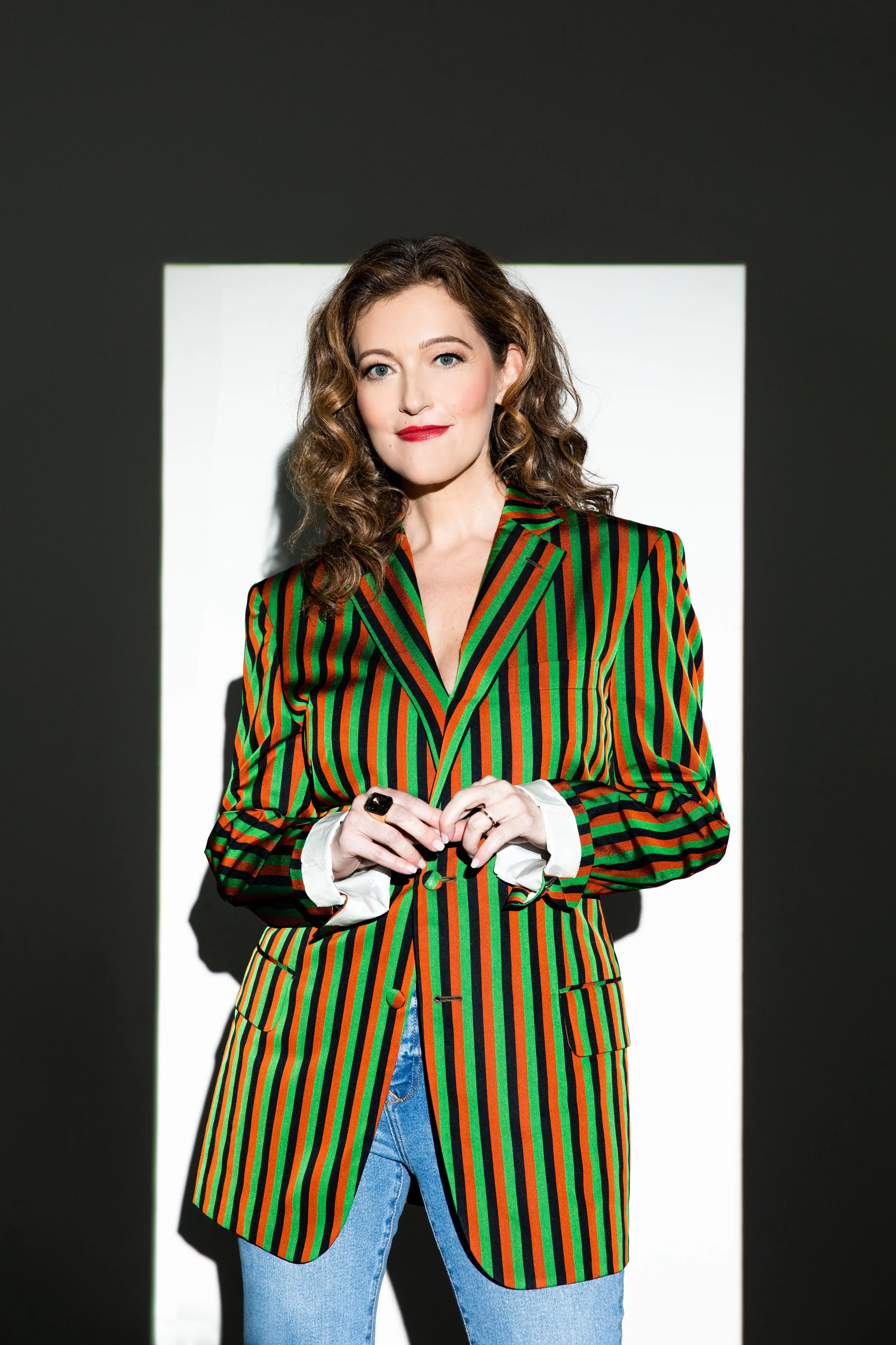 Woman with curly brown hair wearing a colorful striped blazer and blue jeans, standing against a black and white background.