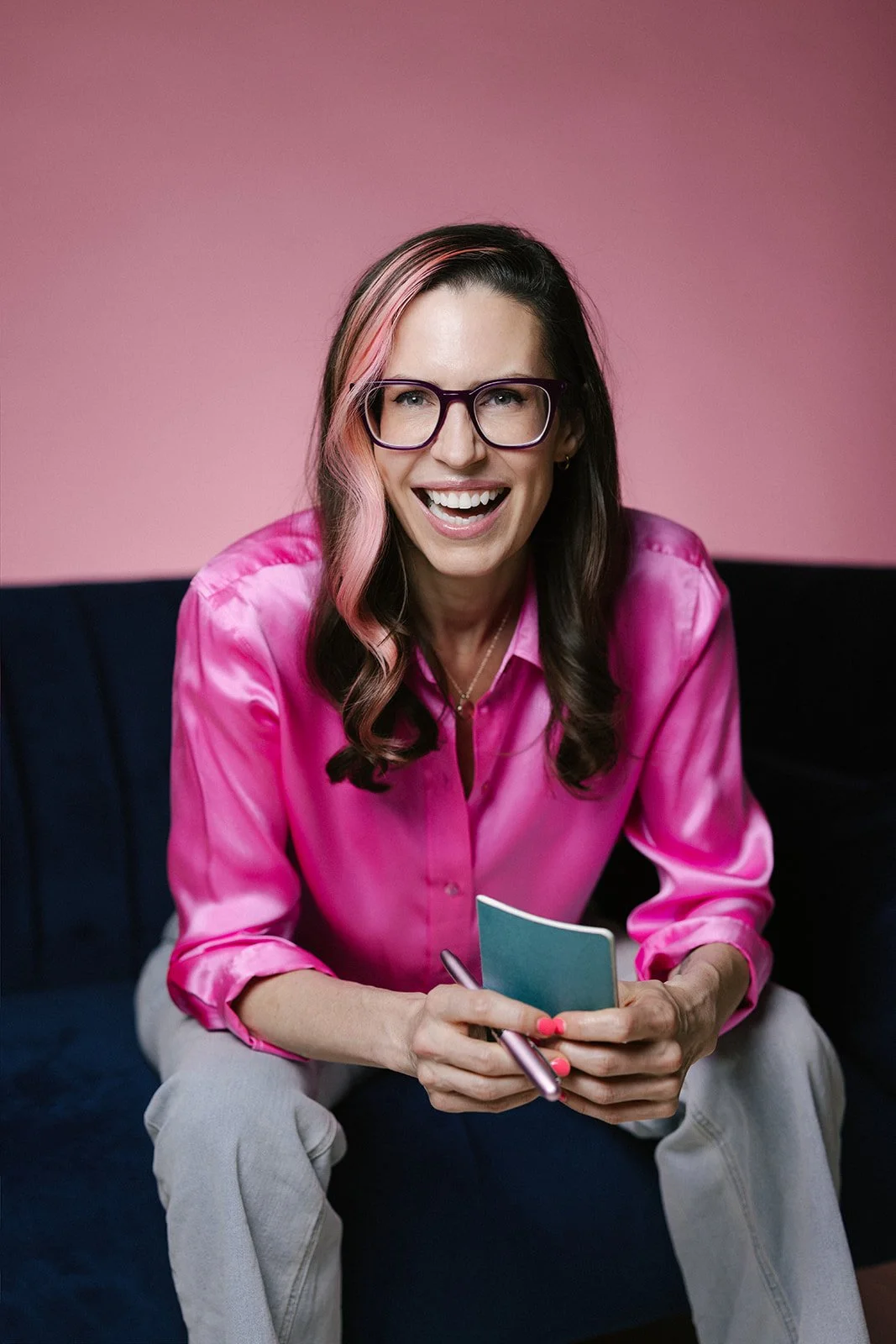 Woman with long dark hair, pink streaks, wearing glasses and a pink satin blouse, sitting on a dark couch, smiling, holding a notepad and pen against a pink wall background.