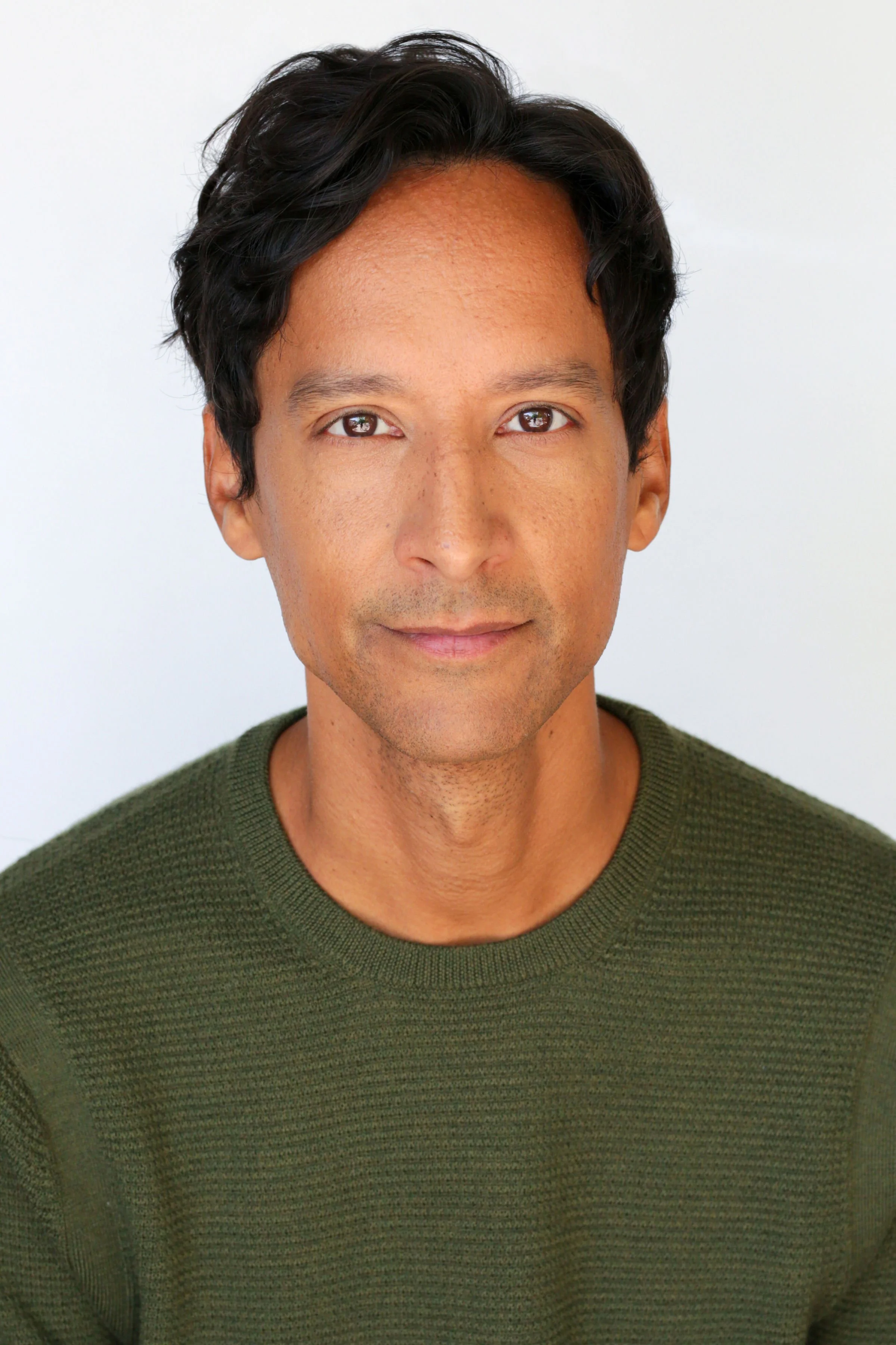Close-up of a young man with dark hair and brown eyes, wearing a green sweater, smiling softly against a plain white background.