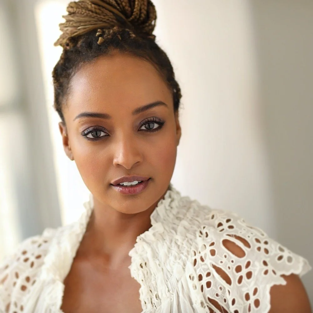 A woman with brown skin, styled in an updo with dreadlocks, wearing a white lace top, is looking at the camera inside a bright room.