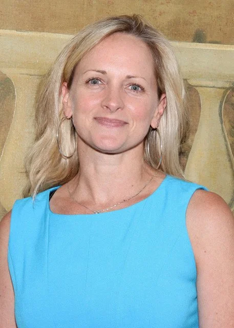 A woman with shoulder-length blonde hair, wearing a sleeveless blue dress and large hoop earrings, smiling at the camera against a blurred background of a wall with decorative pattern.