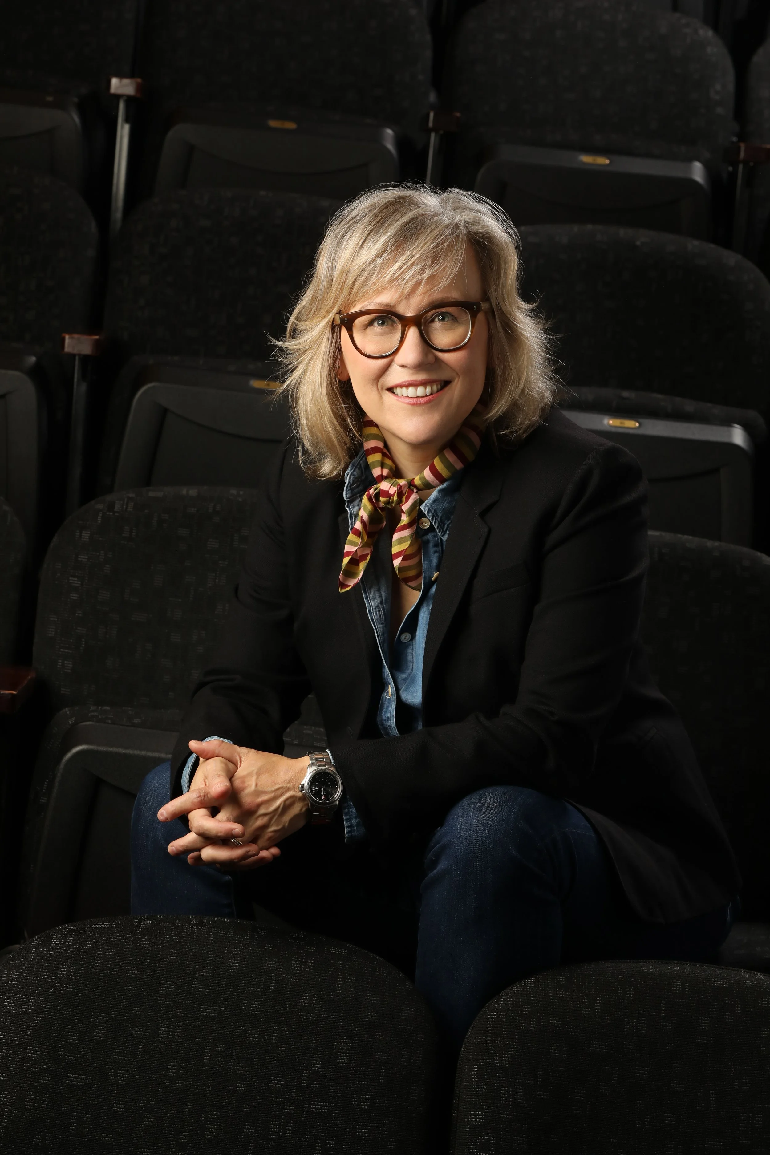 A woman with blonde hair, glasses, and a colorful scarf sitting in a dark auditorium with empty black seats, smiling at the camera.