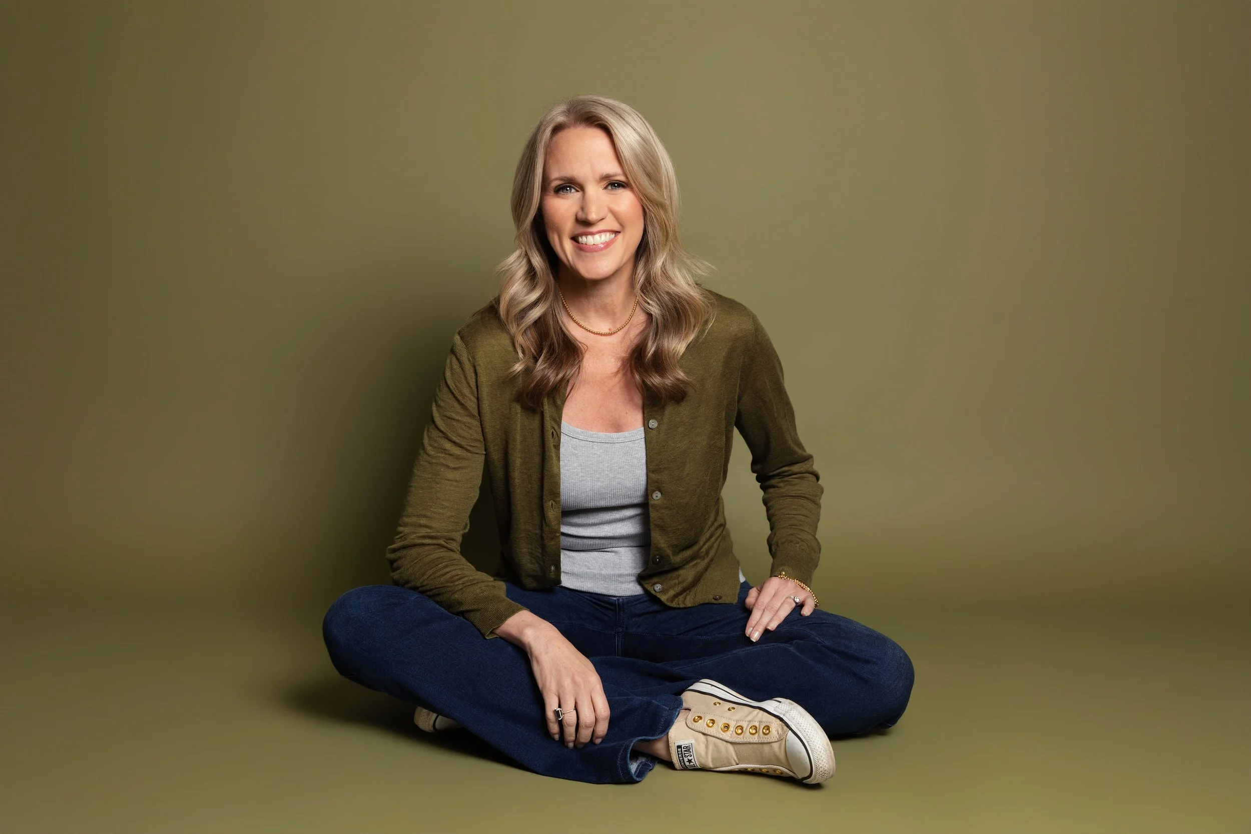 A woman with blonde hair sitting on the floor against a plain olive-green background, smiling and looking at the camera. She is wearing a green cardigan, a gray top, dark jeans, and beige sneakers, with a gold necklace.