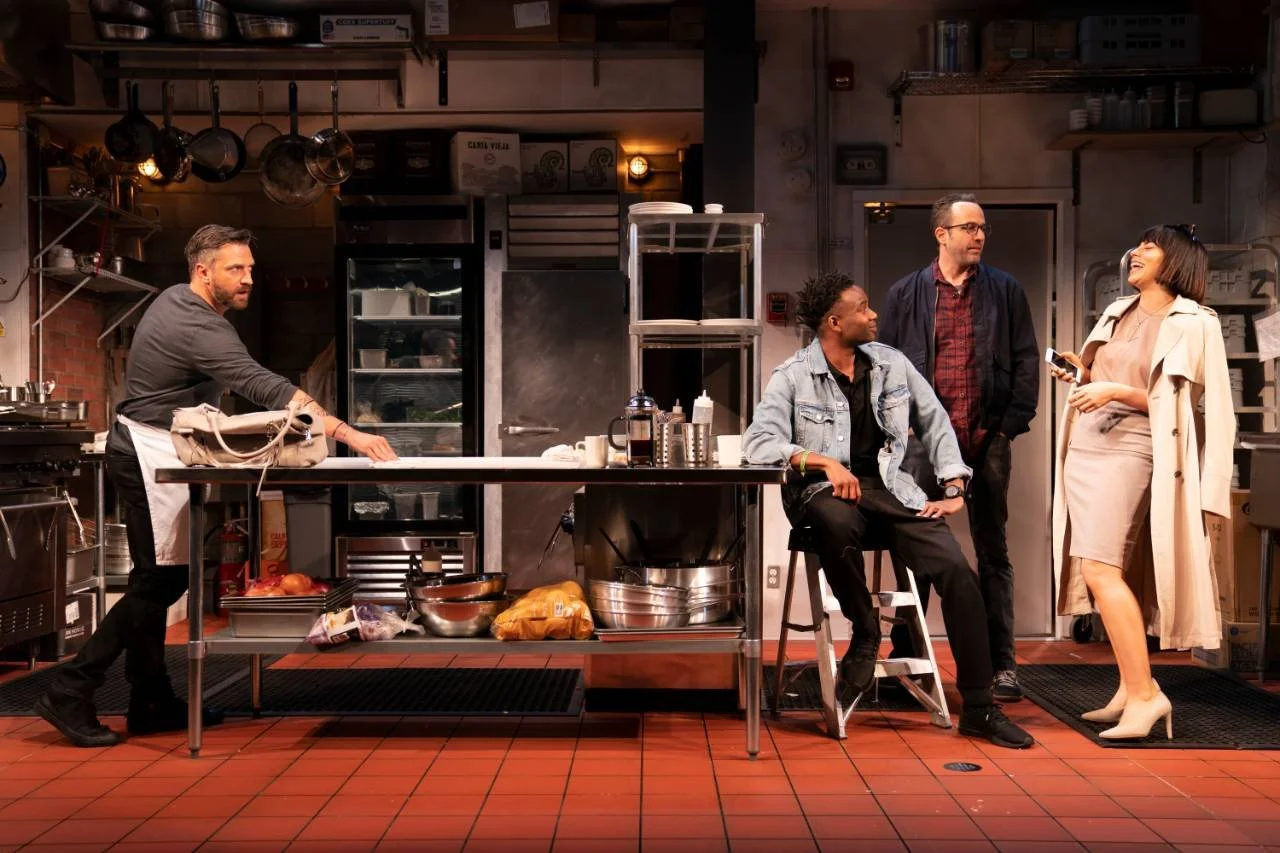 Four people in a restaurant or kitchen setting, engaged in conversation. One man is behind a counter, another is seated on a stool, and two others are standing, woman holding a phone.