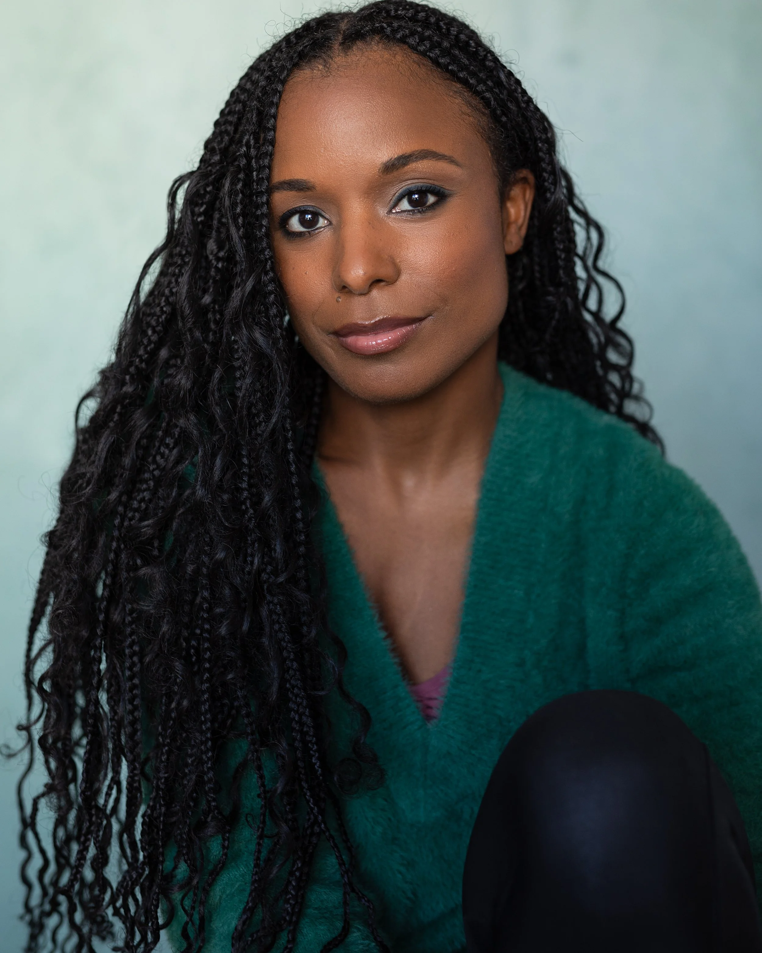 Close-up portrait of an African American woman with long braids, wearing a green sweater, looking softly at the camera with a neutral expression against a light background.