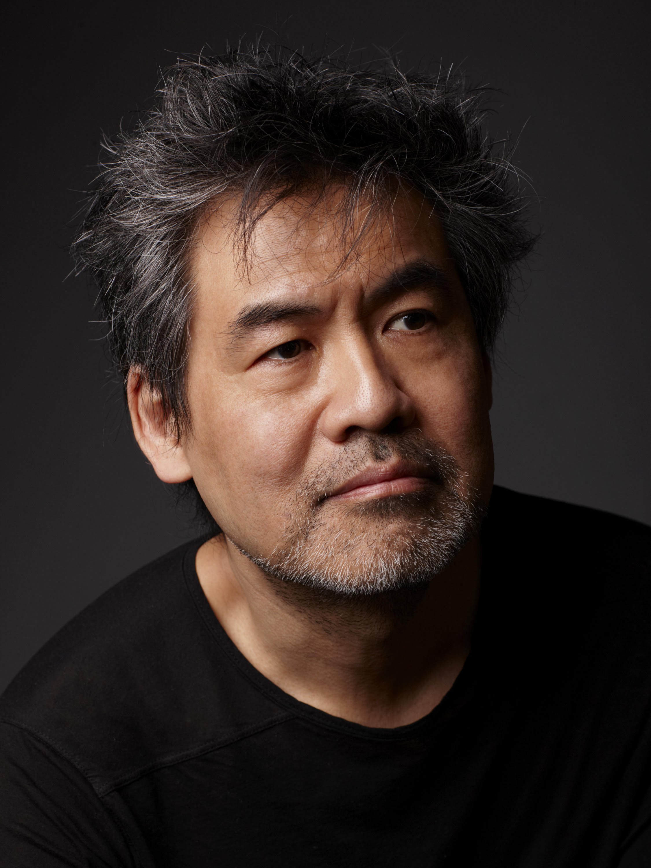 Close-up of a middle-aged man with graying hair and facial hair, wearing a black shirt, against a dark background.