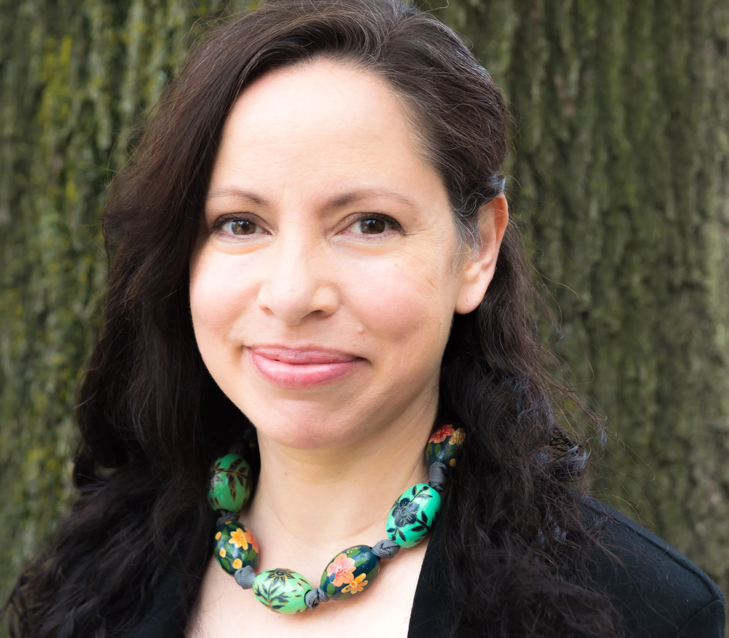 Close-up of a smiling woman with dark wavy hair, wearing a colorful beaded necklace with large green beads and orange flowers, outside with blurred green trees in the background.