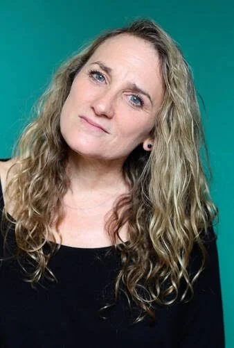 A woman with long curly blonde hair, wearing a black top, is looking at the camera with a slight tilt of her head against a teal background.