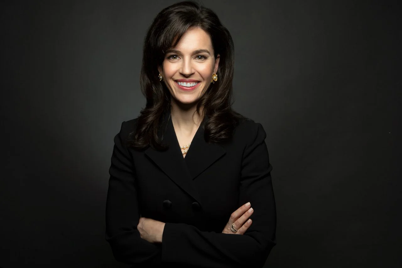 Portrait of a smiling woman with dark brown hair, wearing a black blazer, pearl earrings, and a pearl necklace, against a dark background.