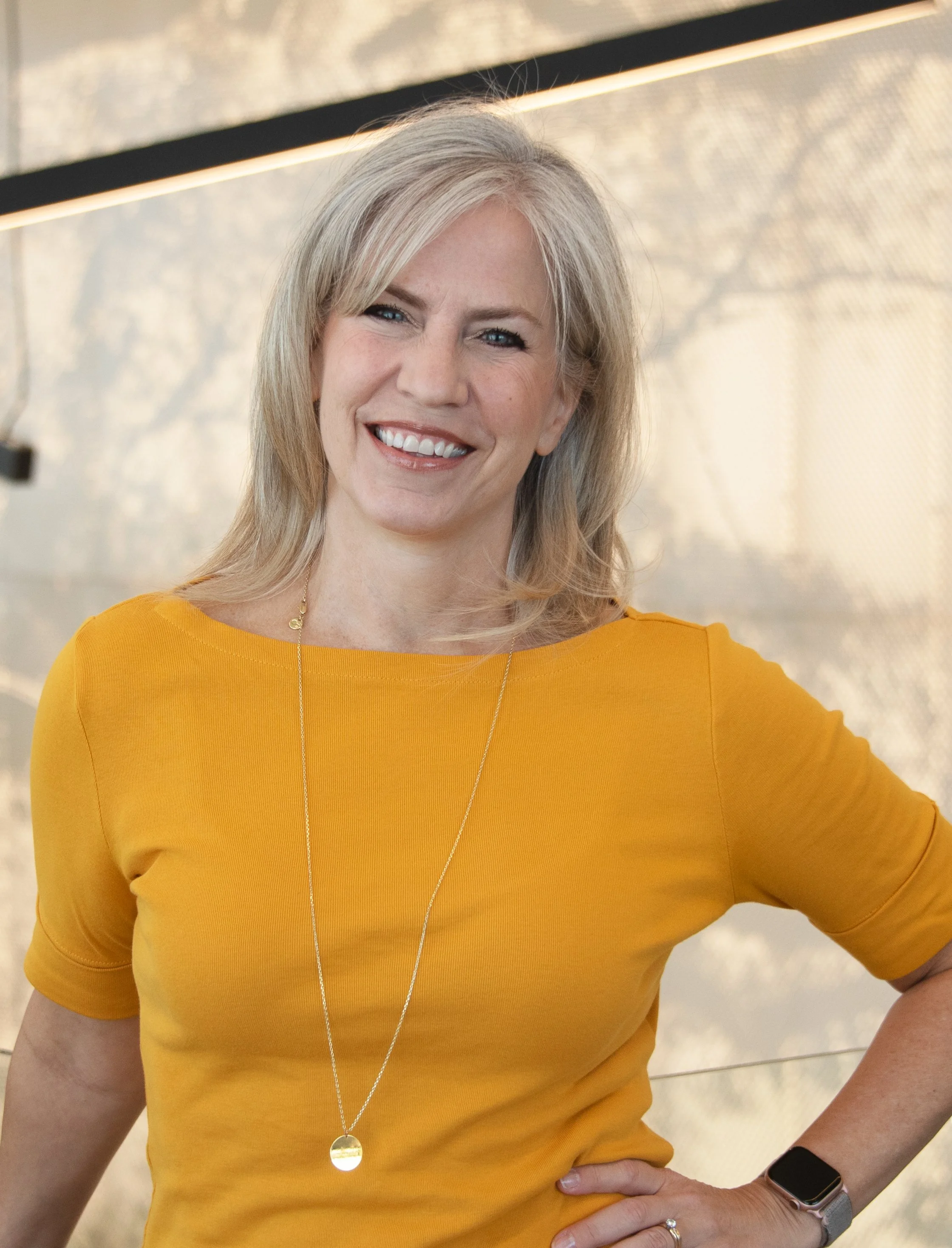 A woman with blonde hair smiling, wearing a yellow top, a gold necklace, a wristwatch, and a ring, standing against a modern interior background.