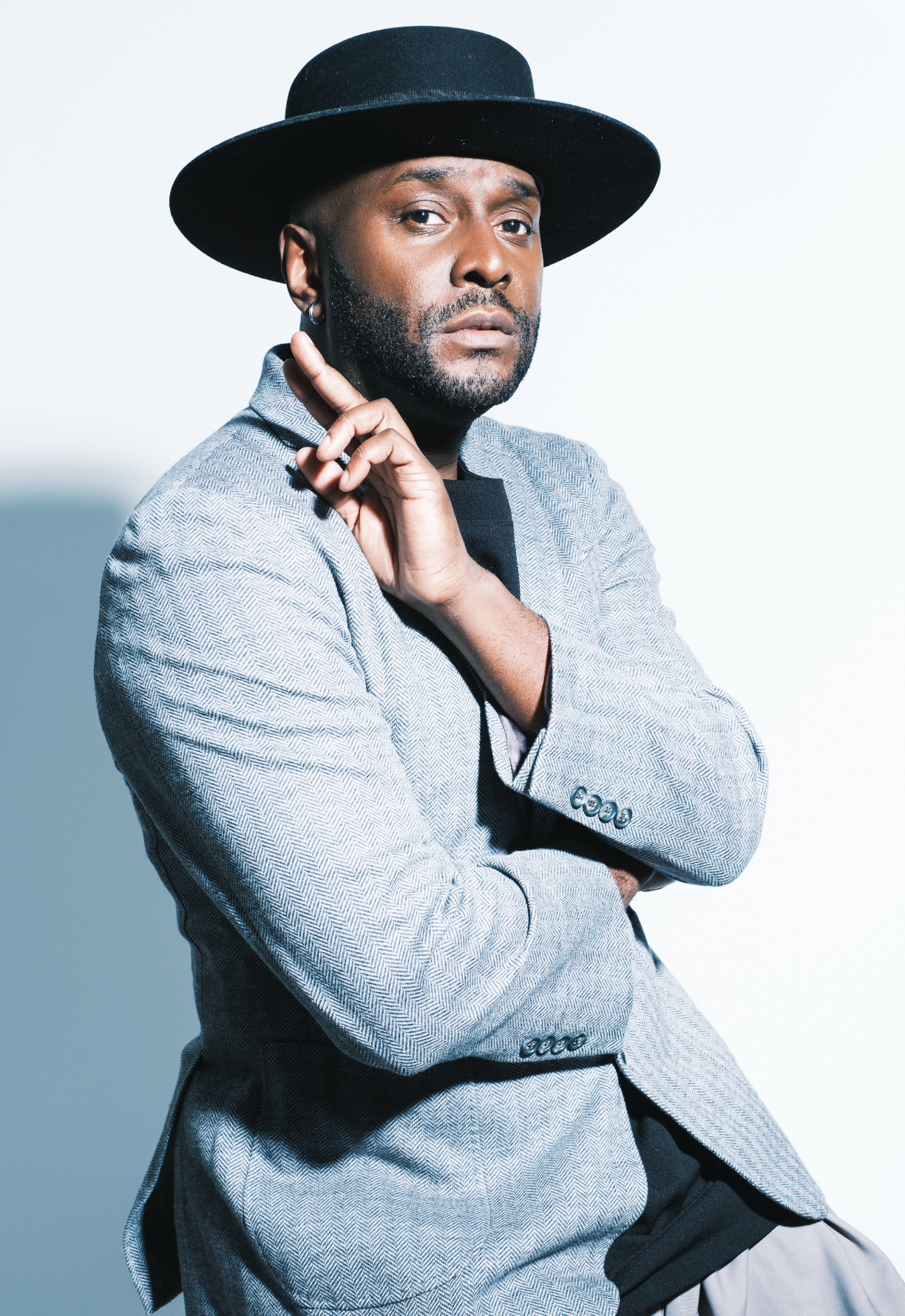 A man wearing a light gray blazer, black shirt, and a black wide-brimmed hat poses against a plain white background.