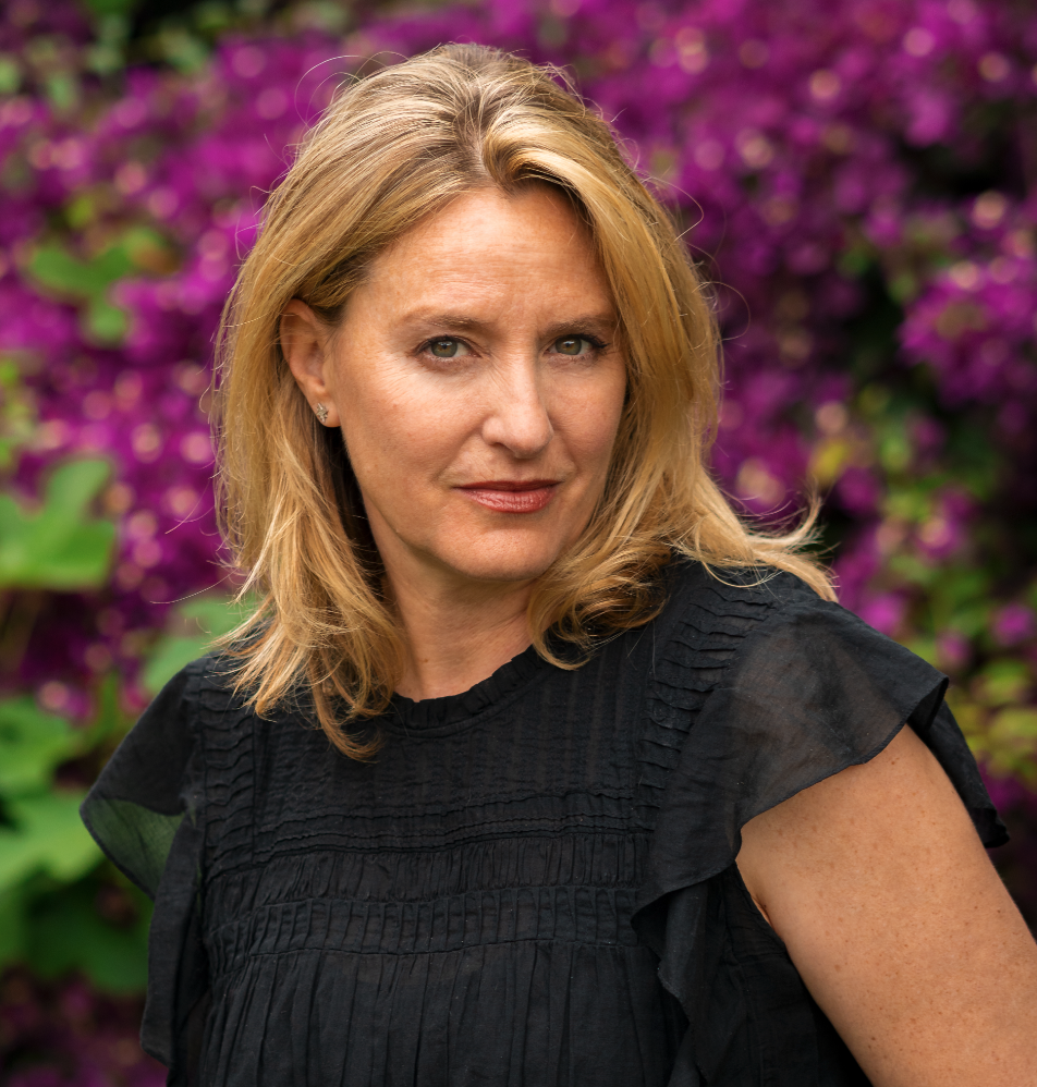 A woman with blonde hair and fair skin posing outdoors in front of purple flowers, wearing a black top.