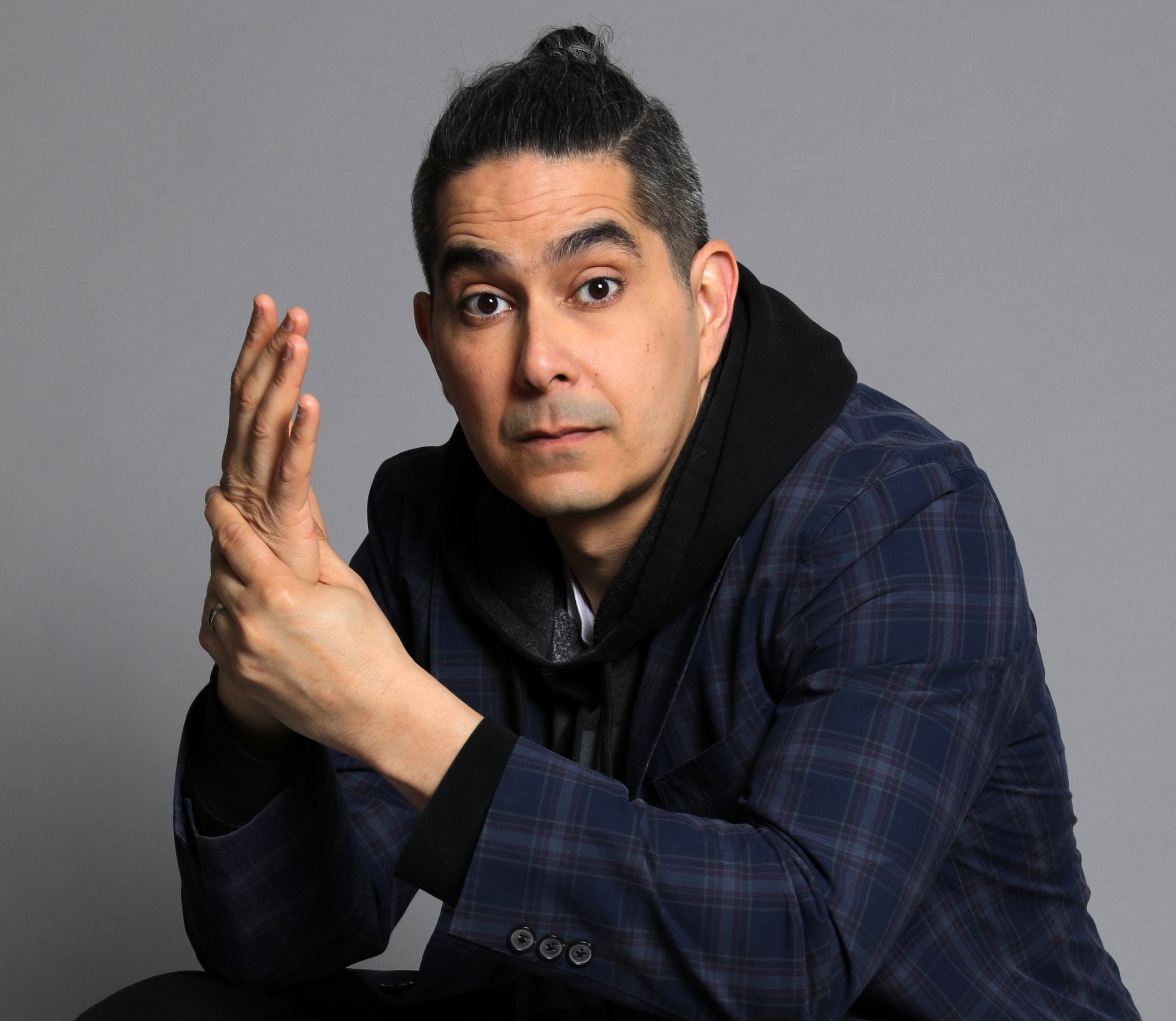 A man with dark hair styled in a bun, wearing a dark plaid blazer and black shirt, posing with his hands near his face against a plain gray background.