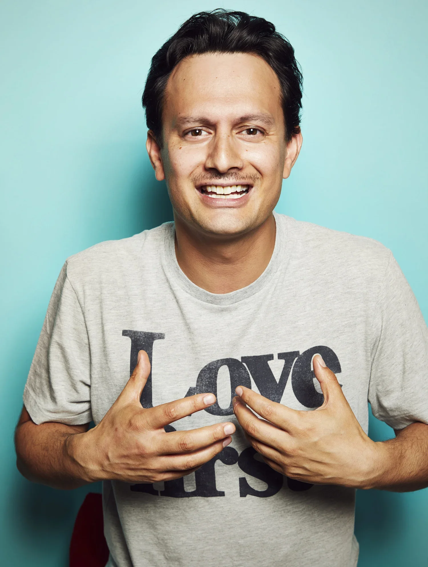 Smiling man with dark hair in a light gray shirt showing love with his hands against a solid blue background.