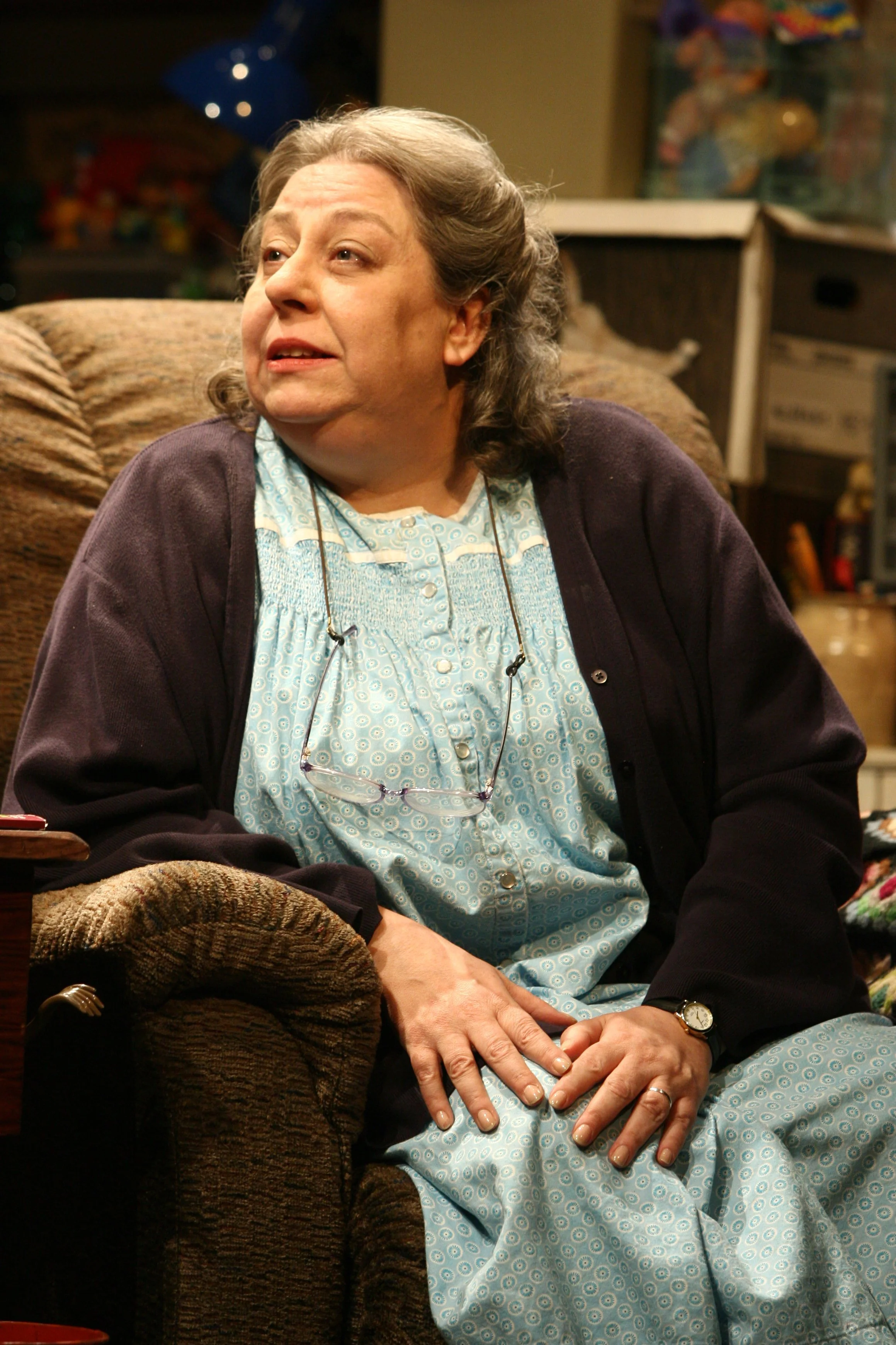 A middle-aged woman with gray hair sitting on a couch in a room with various colorful objects in the background. She is wearing a light blue patterned dress, a dark cardigan, glasses hanging around her neck, and a wristwatch. She appears to be engaged in conversation or thought.