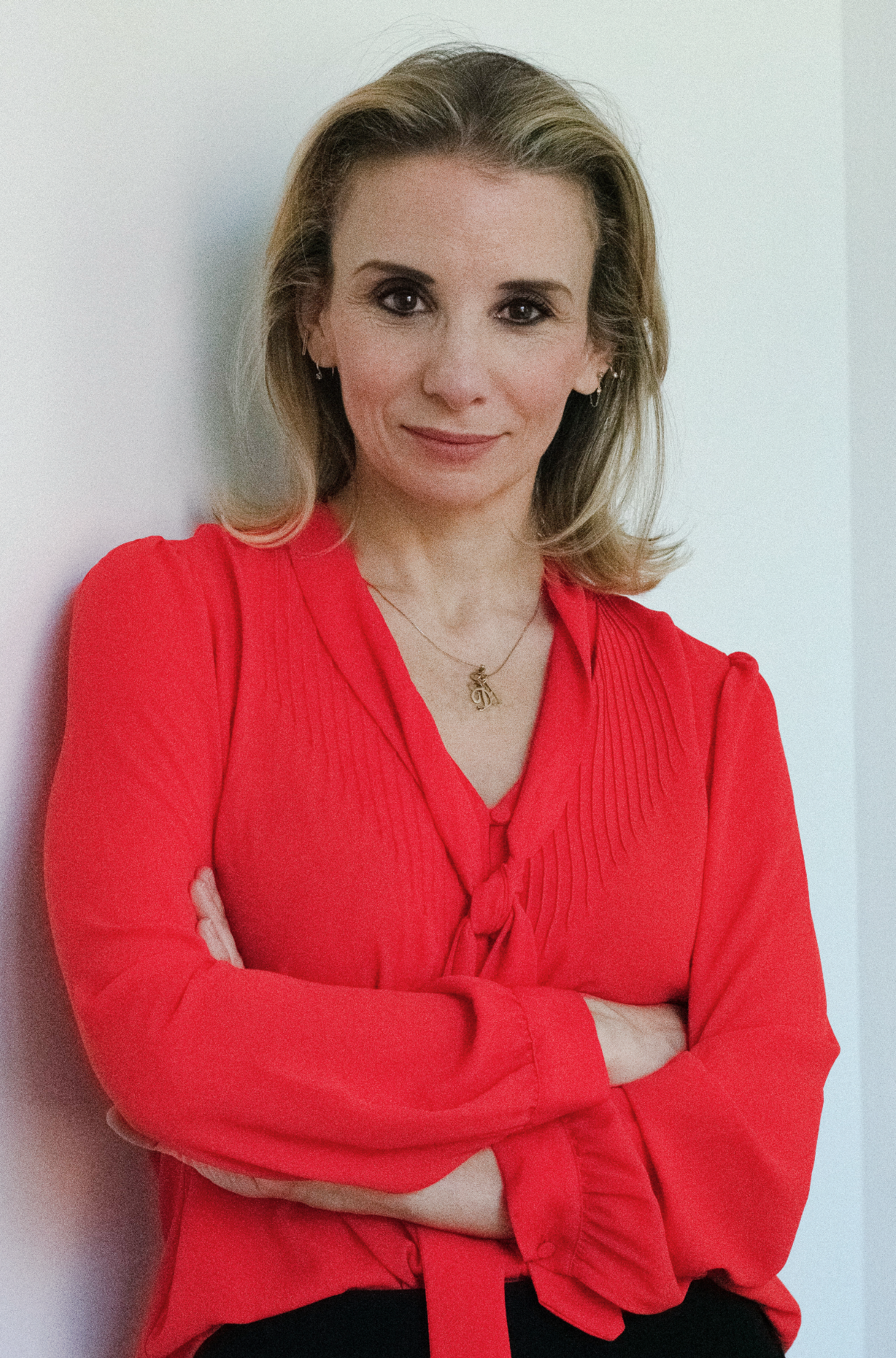 A woman with shoulder-length blonde hair, wearing a red blouse with a tie at the neck, standing against a plain light-colored wall with her arms crossed and smiling slightly.