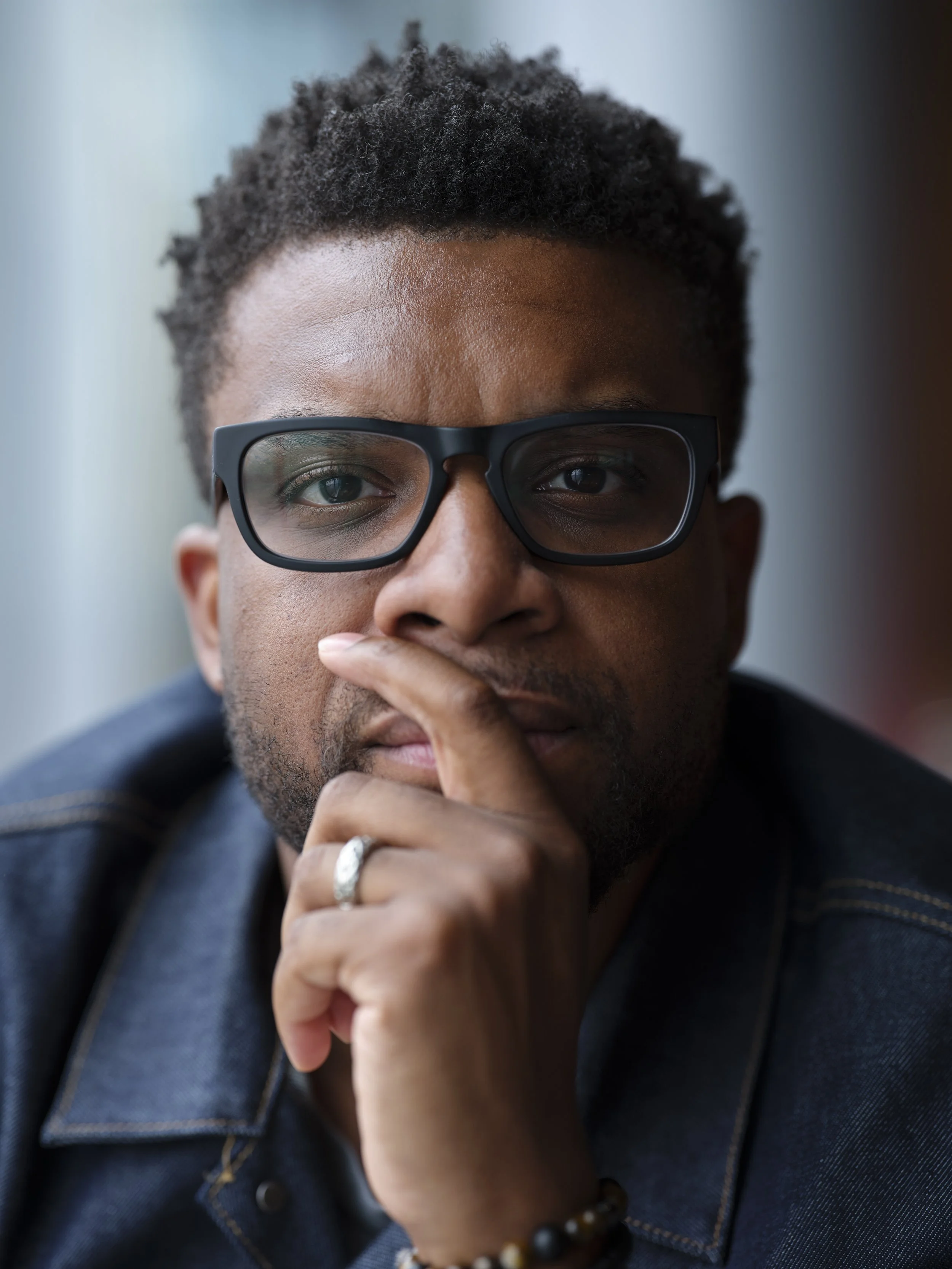 Close-up of a person with short curly hair wearing glasses, touching their chin with one hand, wearing a dark jacket and a beaded bracelet.