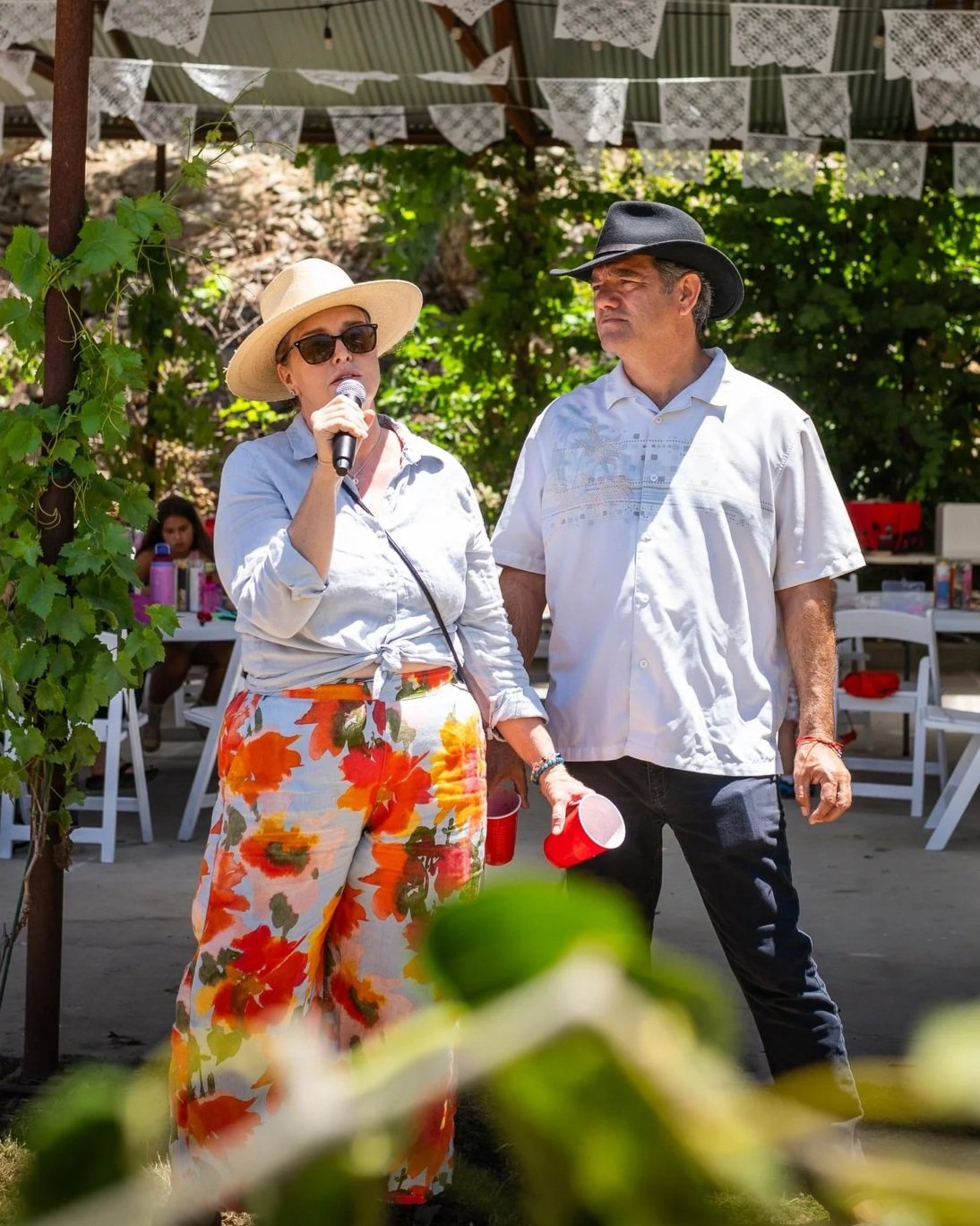 A woman wearing a wide-brimmed hat, sunglasses, and floral pants is holding a microphone and speaking. A man wearing a black cowboy hat, white shirt, and dark pants is standing next to her. They are outdoors under a decorated canopy, with greenery and tables in the background.