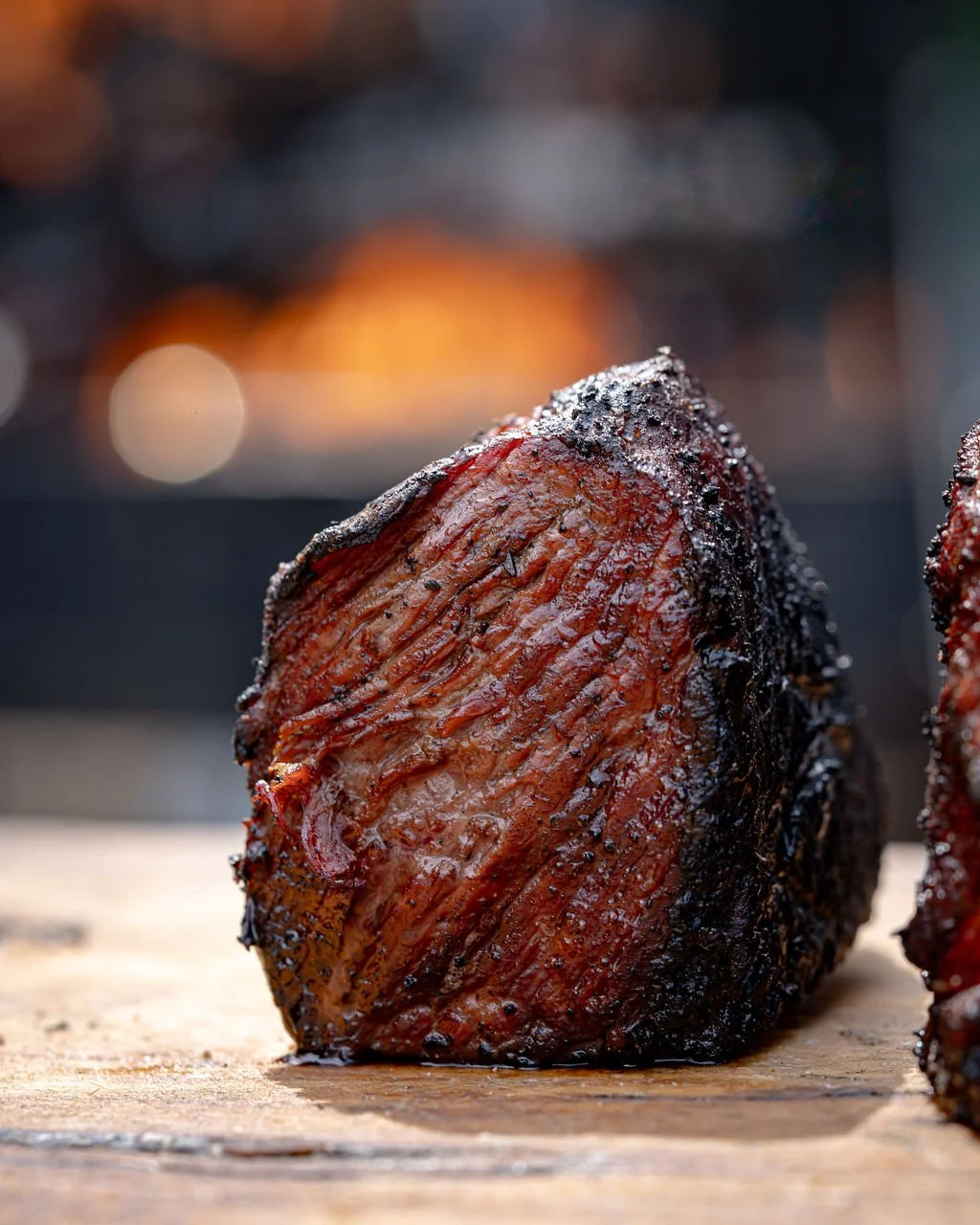 Close-up of a slice of smoked or grilled brisket on a wooden surface, with a blurred background.