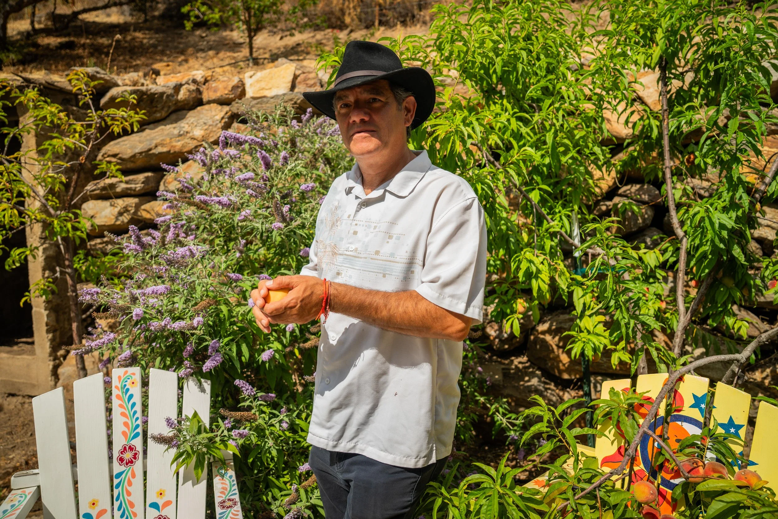 A man wearing a black cowboy hat and a white button-up shirt stands outdoors near a garden with purple flowers and green foliage, holding a yellow object in his hands.