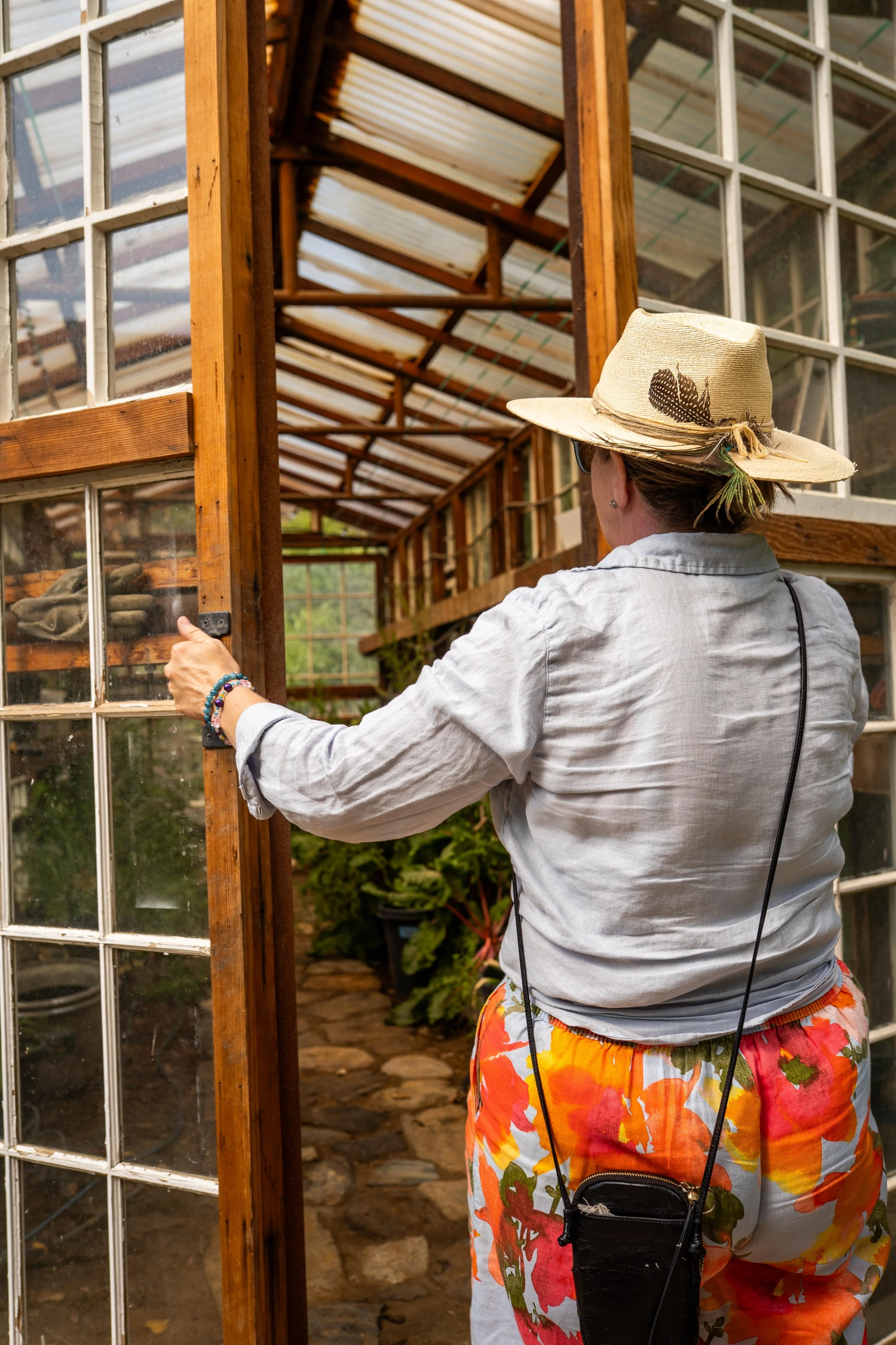 A woman wearing a straw hat with a feather, sunglasses, a light gray shirt, and colorful floral shorts, opening the door of a greenhouse.