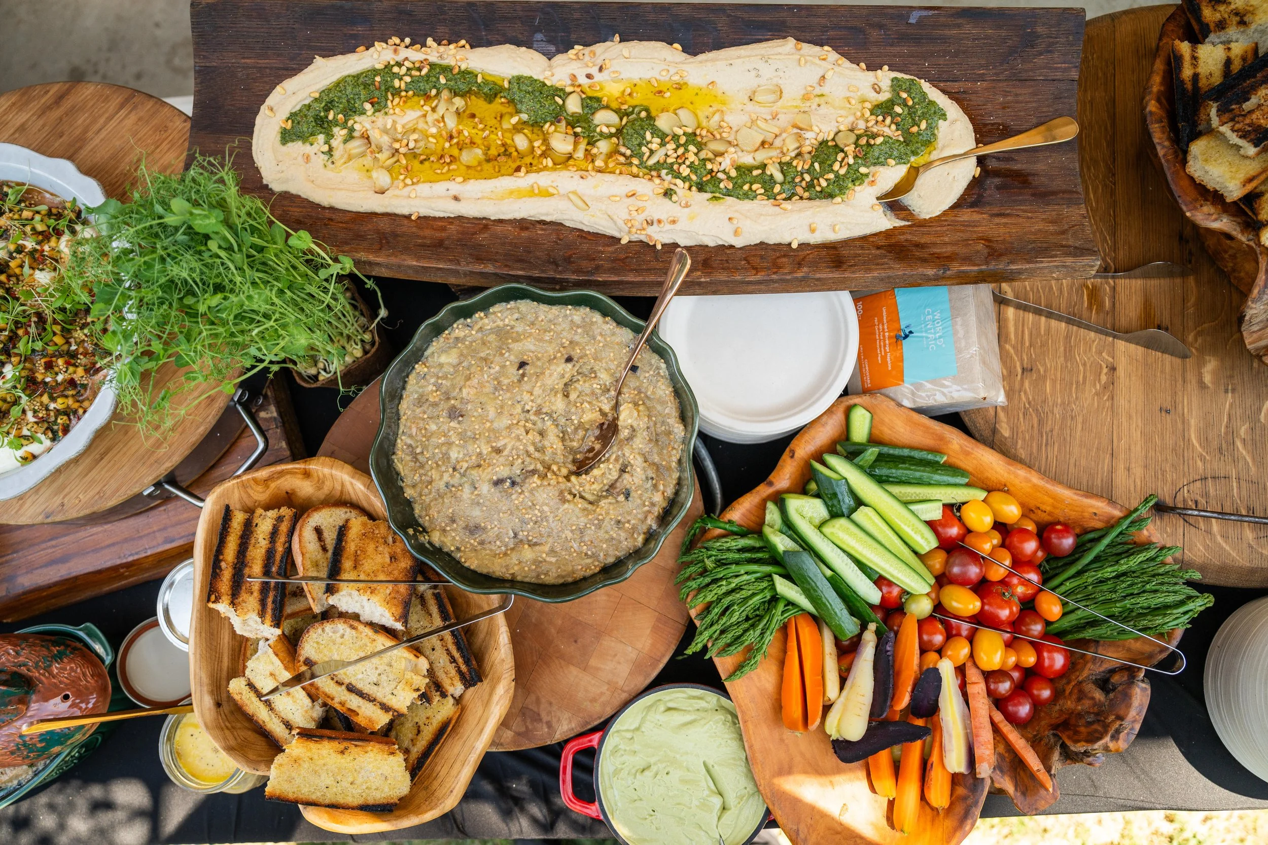 Assorted colorful vegetables, dips, bread, and a large baked flatbread topped with pesto, pine nuts, and olive oil on a wooden platter, part of a buffet or meal spread.