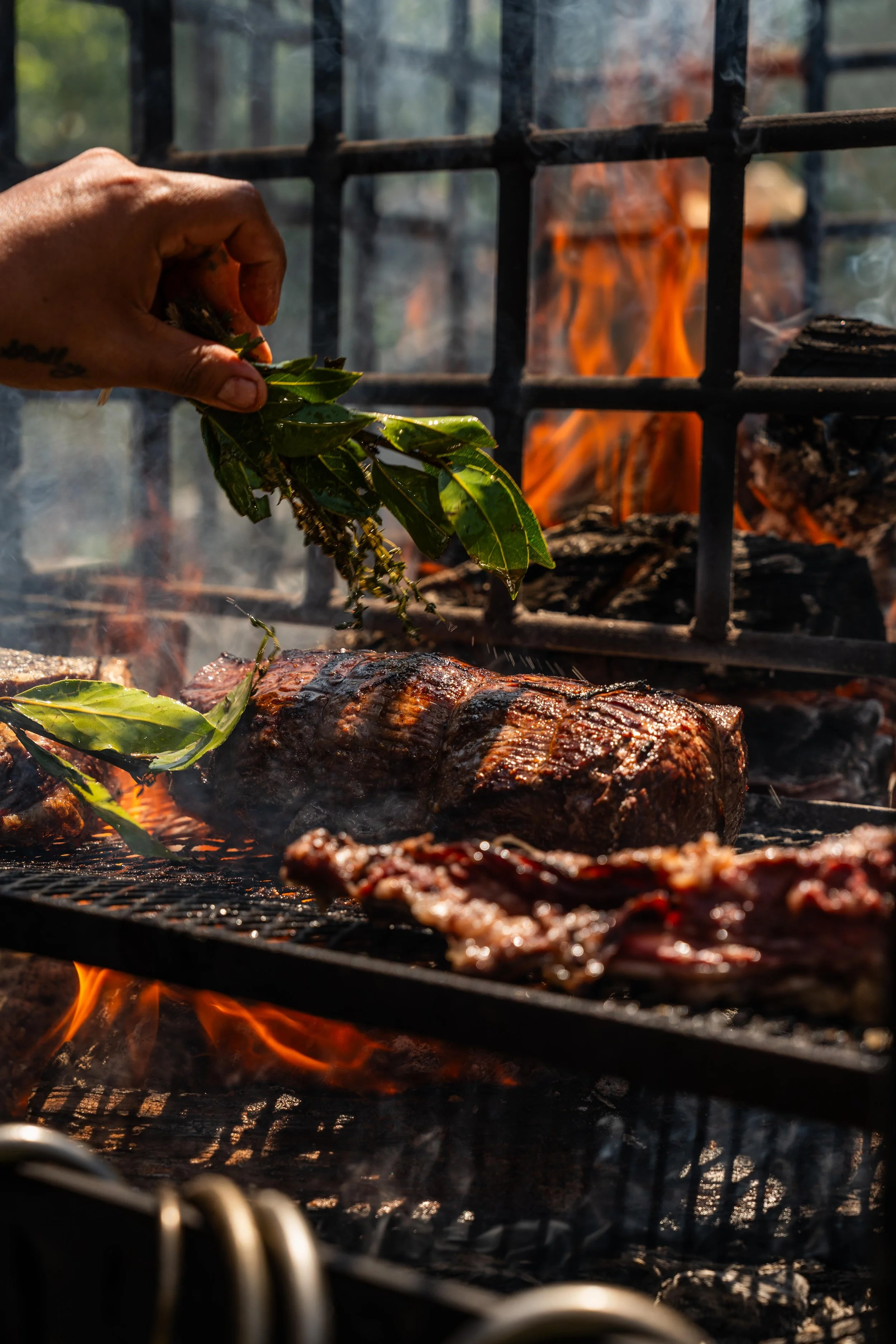 A hand holding fresh herbs over a grill with steak and sausages cooking, fire and flames visible behind the grill.