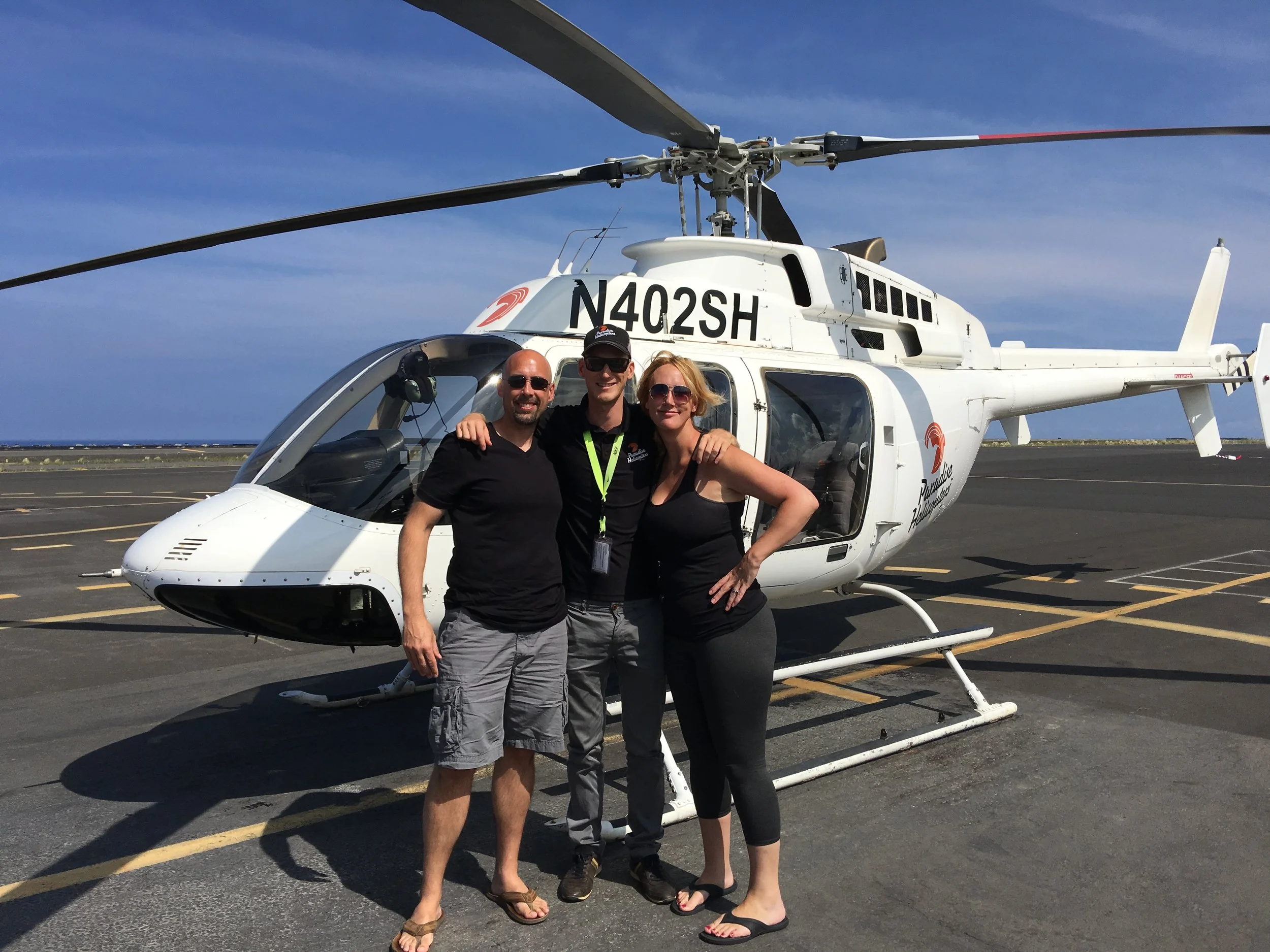 Three people, two men and a woman, standing in front of a white helicopter with registration N402SH on an airport tarmac on a sunny day, smiling at the camera.