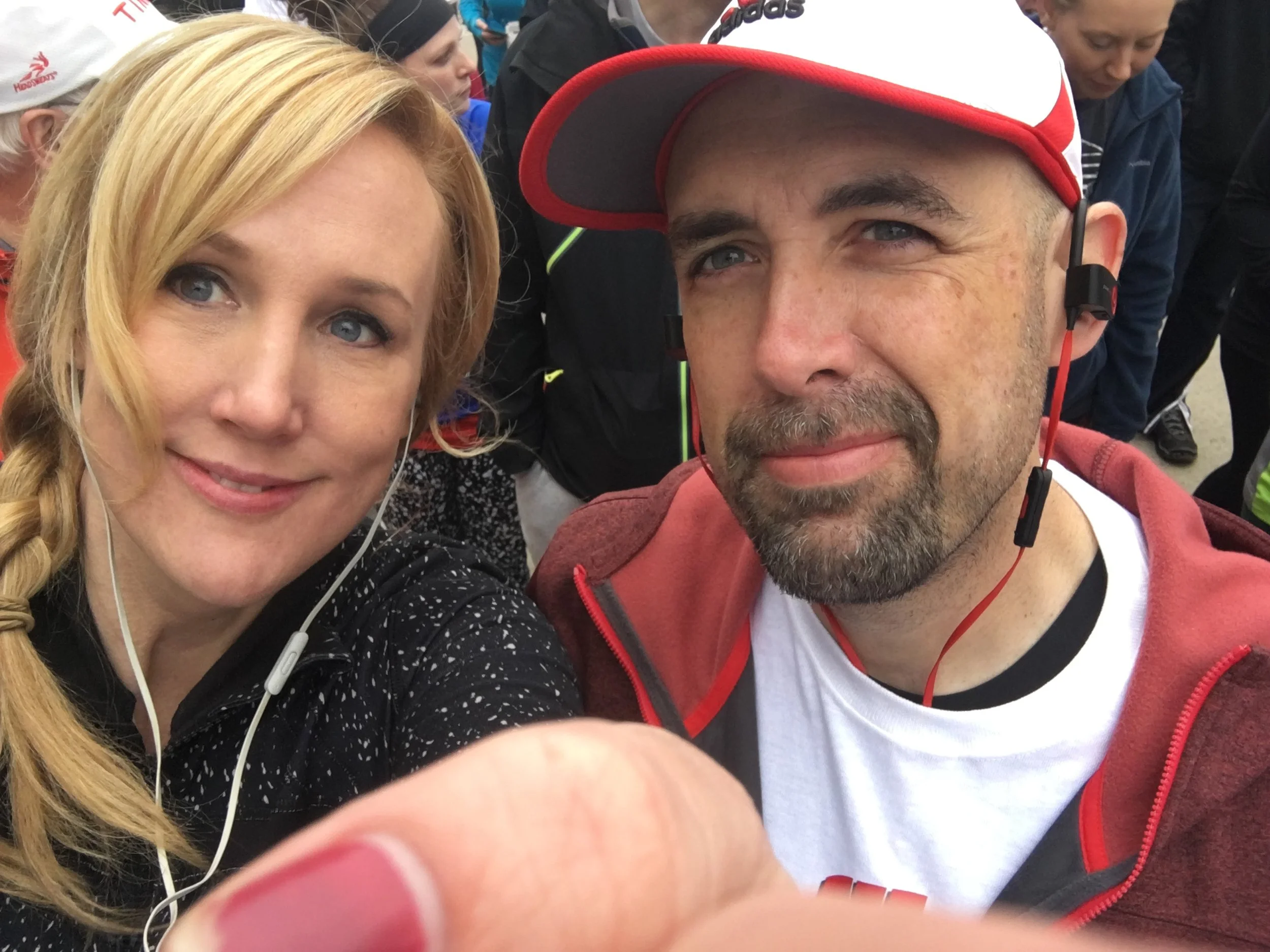 Close-up of a woman with blonde hair and blue eyes and a man wearing a red and white cap, both with earphones, taking a selfie among a crowd of people.