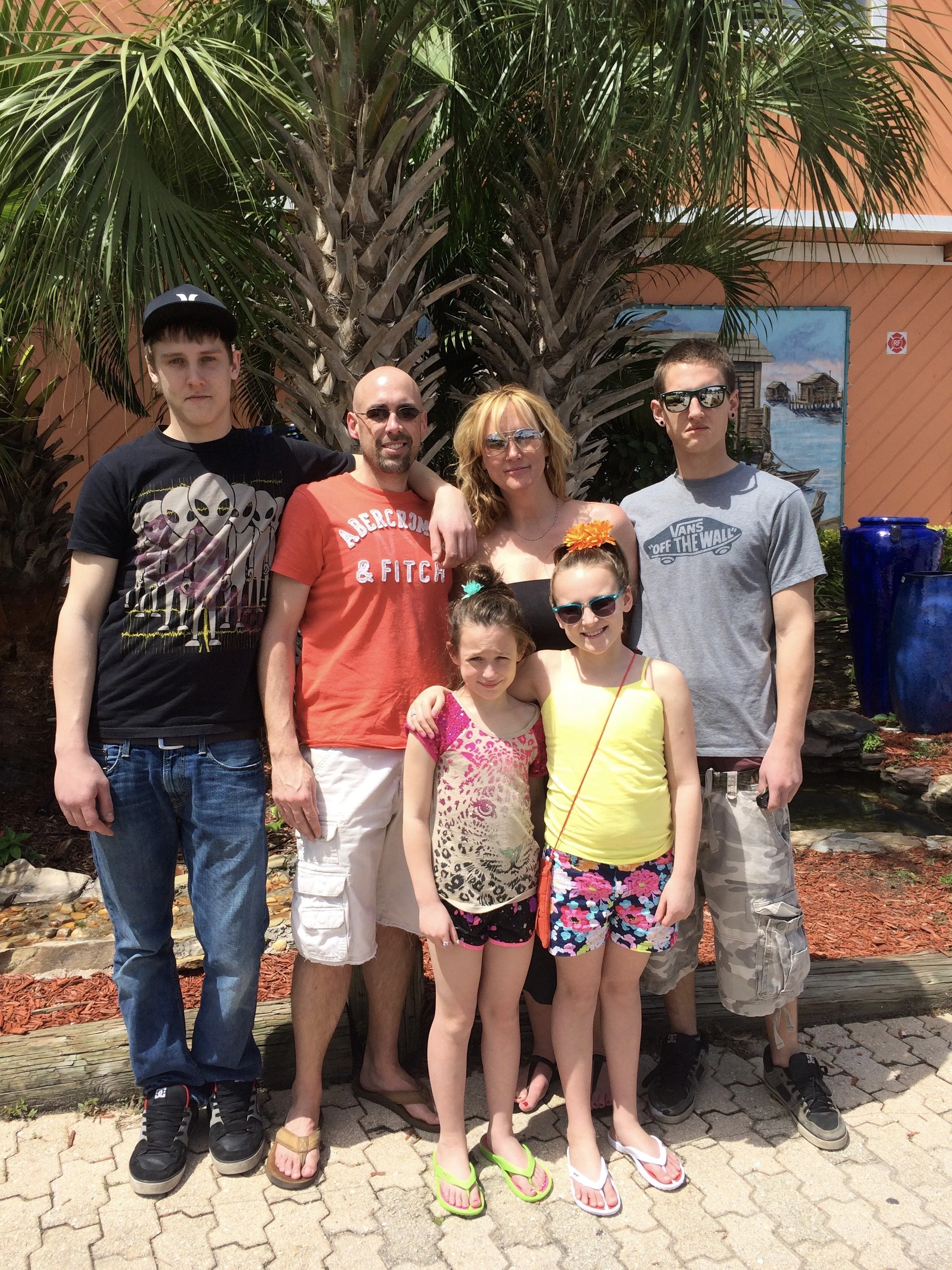 A group of seven people posing outdoors in front of a large palm tree. The group includes three teenagers, a man, a woman, and three young girls. They are standing on a brick paved area, with some wearing sunglasses and casual summer clothing, and sm