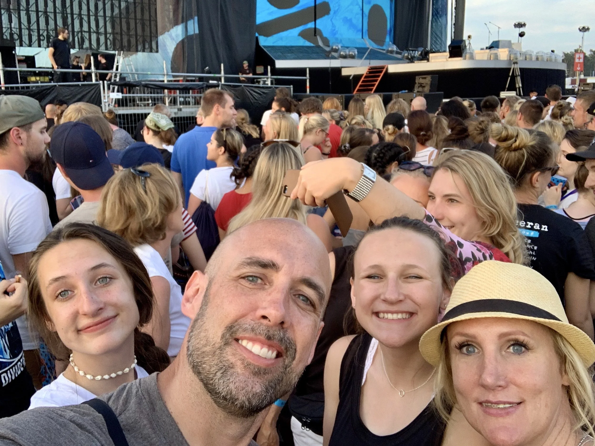 A large group of people, including men, women, and children, gathered outdoors at a concert or festival, with a stage and equipment visible in the background.