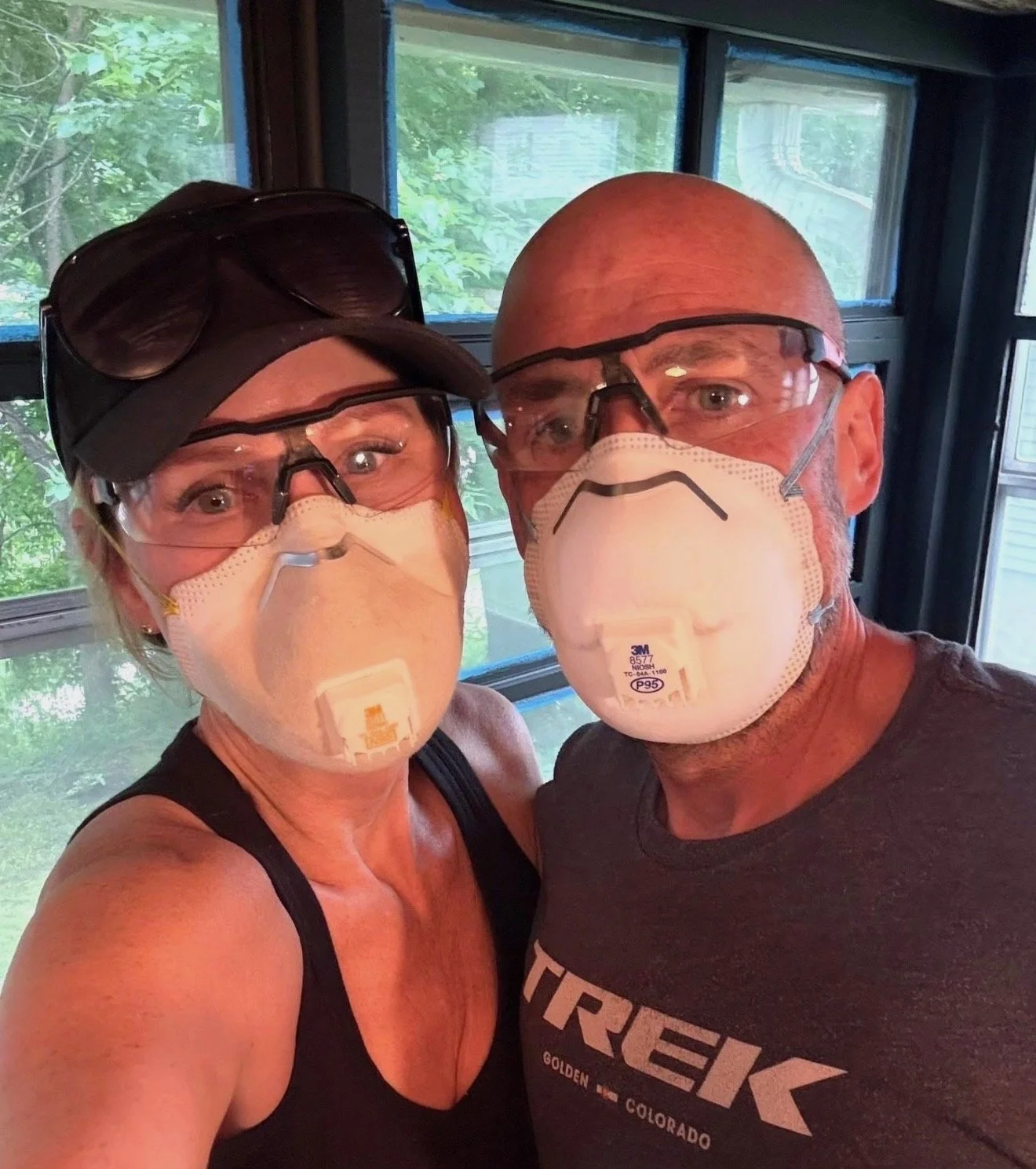 A man and a woman wearing masks and safety glasses, taking a selfie inside a building with large windows showing trees outside.