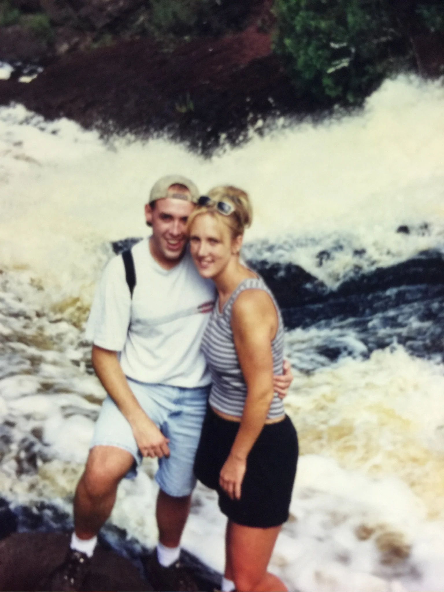 A young man wearing a beige cap, white t-shirt, and shorts, and a young woman wearing sunglasses on her head, a striped tank top, and shorts, are standing close together in front of a turbulent river with white water rapids and rocks, surrounded by l