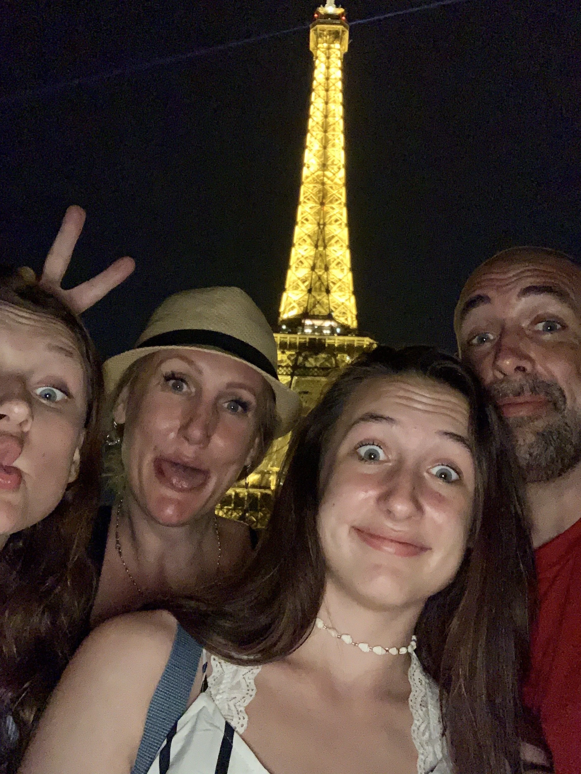 The family taking a selfie at night with the Eiffel Tower illuminated in the background. We are smiling, making silly faces, and one person is giving a peace sign.