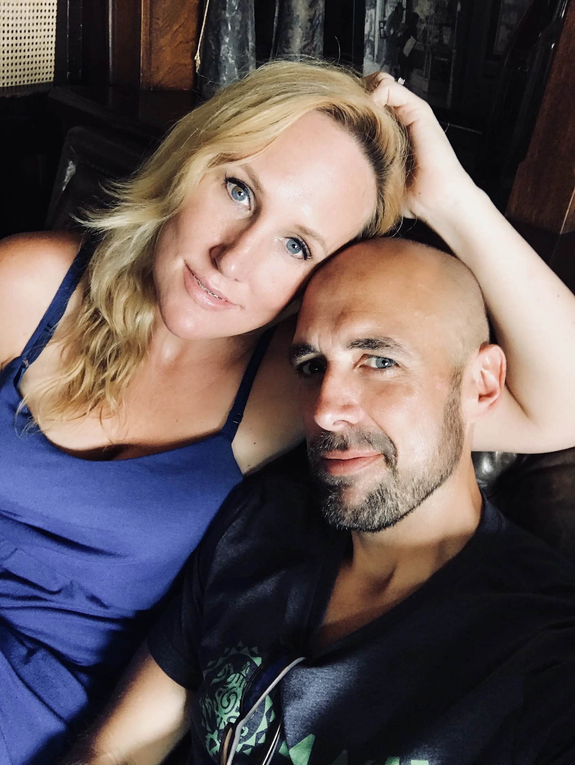 A woman with long blonde hair and blue eyes, and a man with a shaved head and beard, lying close together with the woman's head resting on the man's forehead, indoors with dark wood furniture and a marbled curtain in the background.