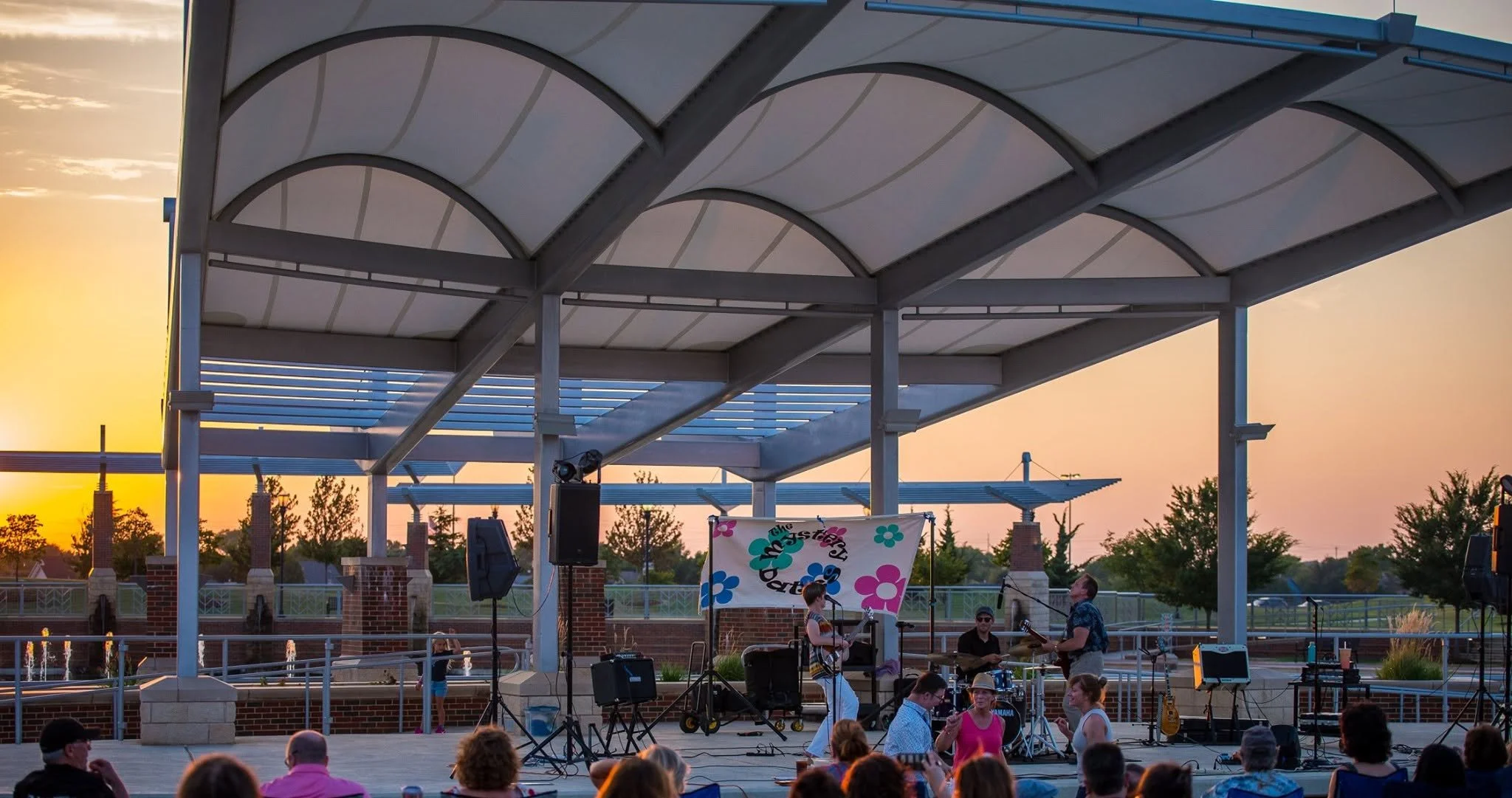 A live outdoor music performance at sunset, with a band playing under a modern white canopy, and an audience watching.