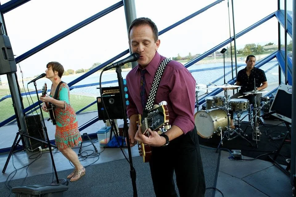 Band performing on an indoor stage with large windows showing an outdoor view; a woman singing and playing guitar, a man singing and playing guitar, and a drummer playing the drums.