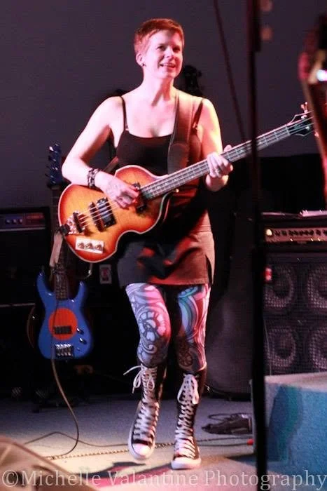 Young woman performing on stage with a guitar, wearing colorful patterned leggings and Converse sneakers.