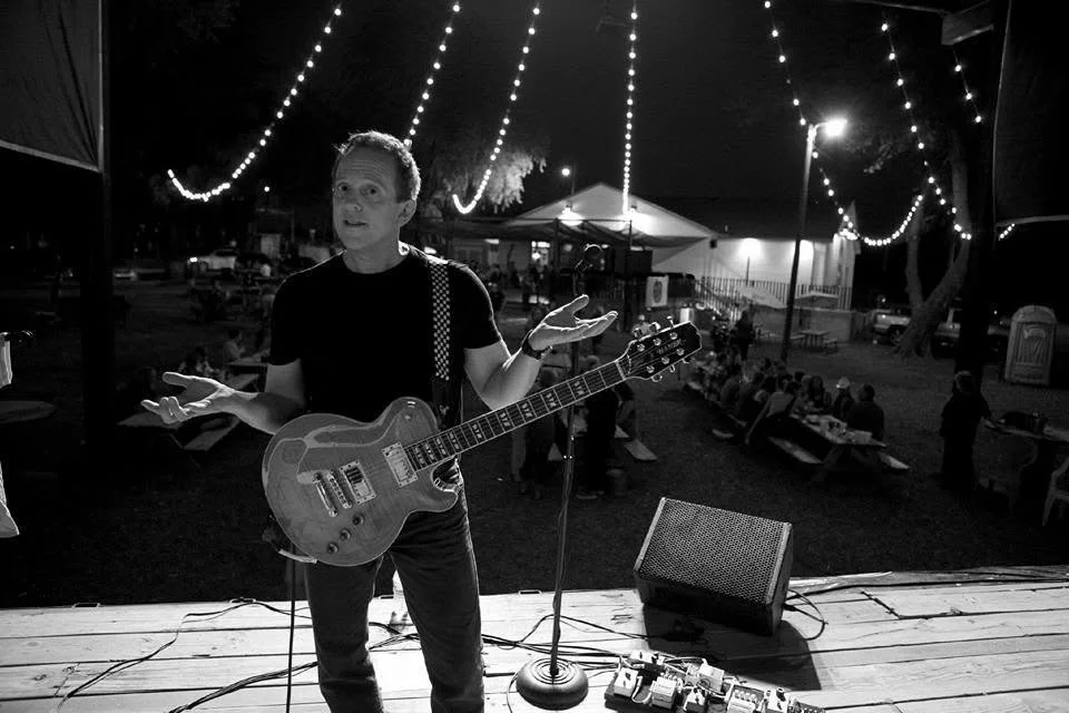 A person standing on an outdoor stage at night, holding a guitar and shrugging with a confused or questioning expression, with string lights overhead and an audience seated at picnic tables in the background.